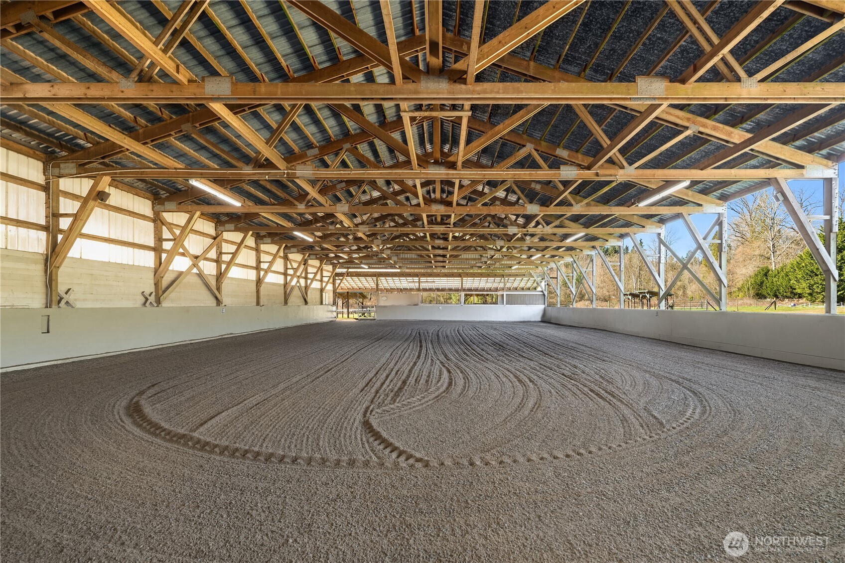 Empty indoor riding arena with groomed footing, exposed timber trusses, and open sides.