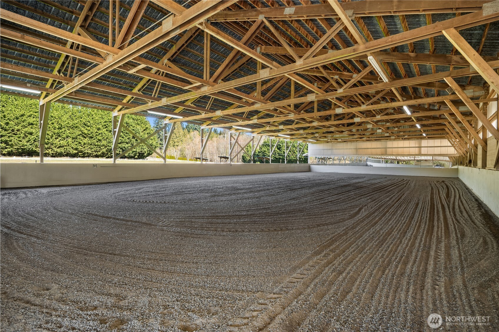 Covered riding arena with a groomed surface and exposed timber roof trusses.