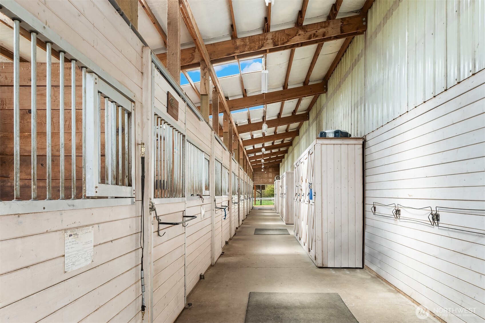 Horse stable aisle lined with white stalls, metal bars, and wooden storage cabinets.