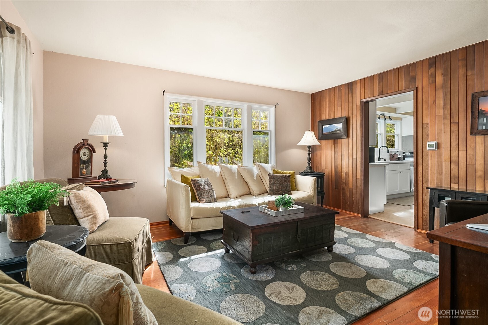 Furnished living room with a wood-paneled accent wall, cream sofas, and large sunlit windows.