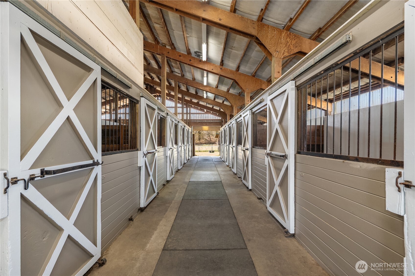Horse stable aisle lined with X-braced stall doors beneath exposed timber roof beams.