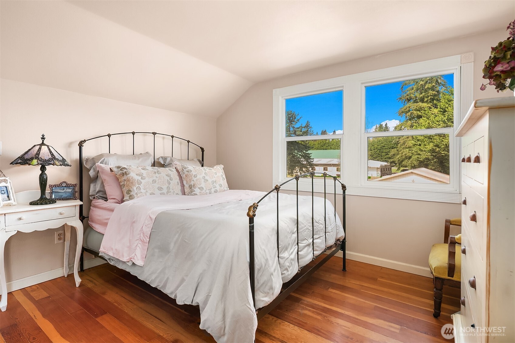 Furnished bedroom with a black metal bed, hardwood floors, and two large windows overlooking trees.