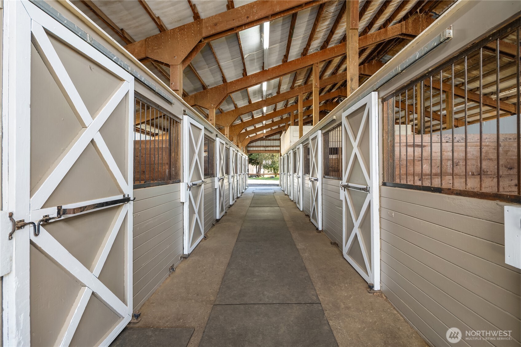 Horse stable aisle lined with gray-and-white stalls beneath exposed timber roof beams.