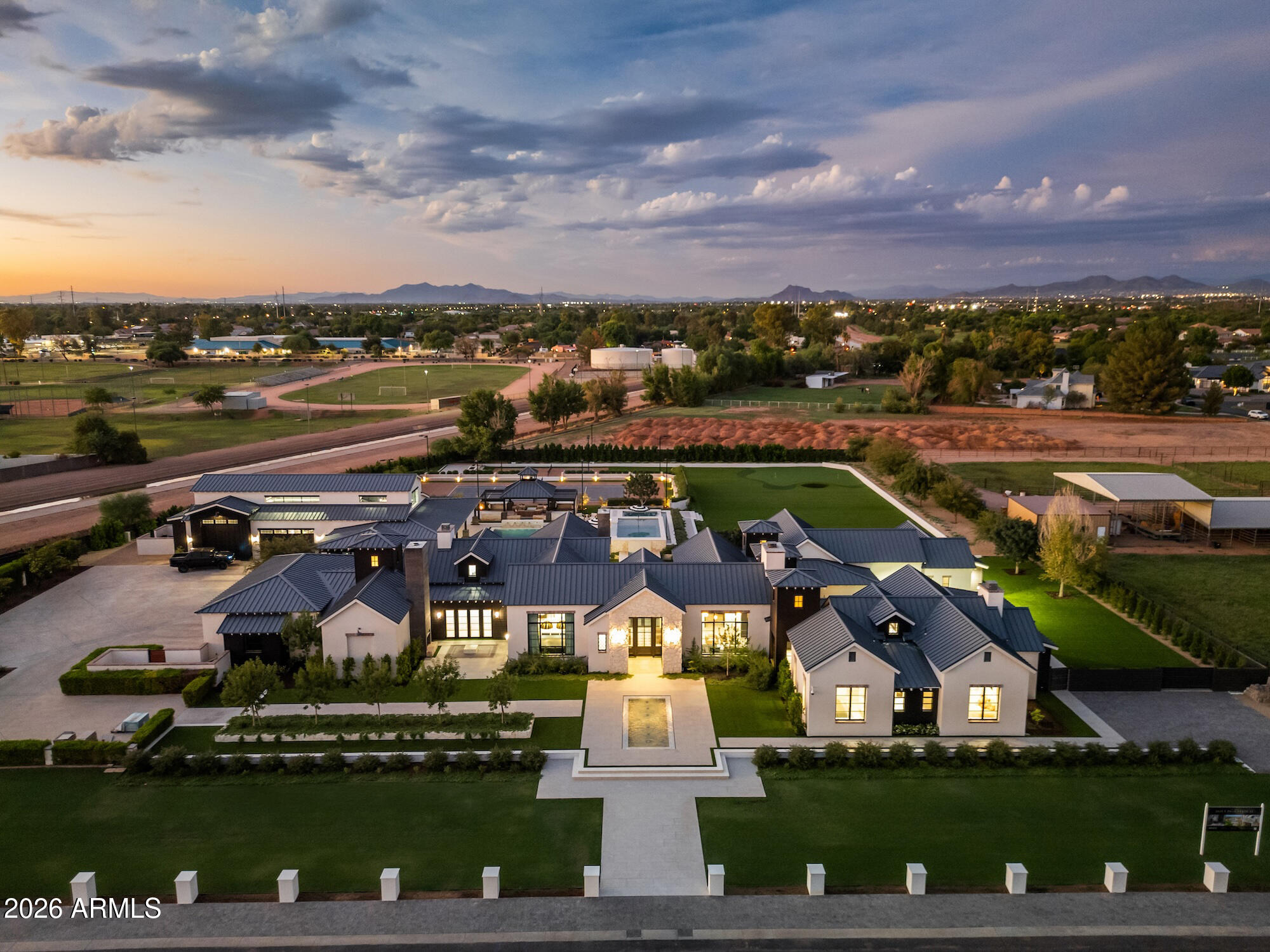 Large luxury home with dark metal roofs, broad lawns, and a pool, photographed from above at dusk.
