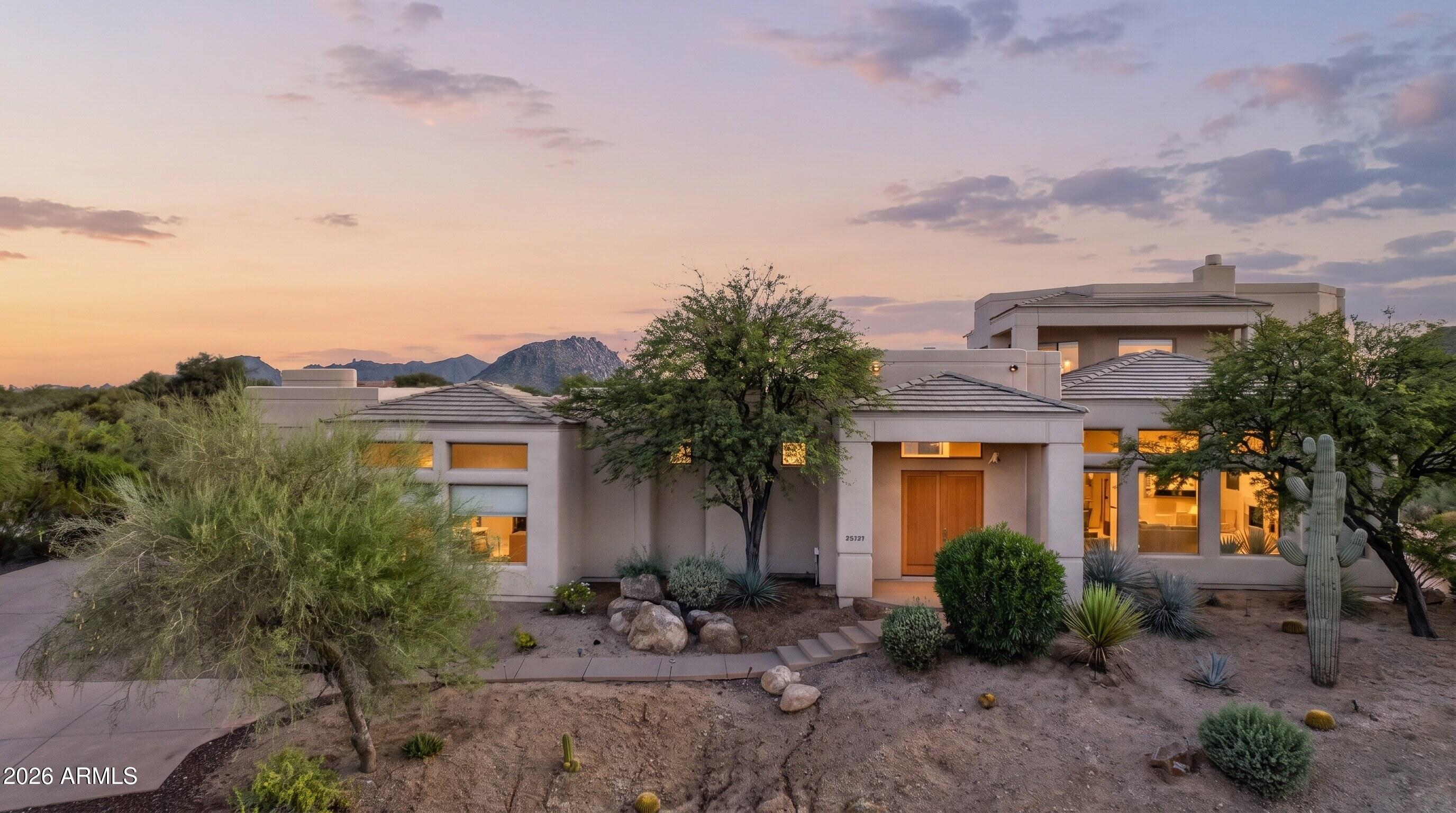 Modern desert home with glowing windows, cacti, and mountains at dusk.