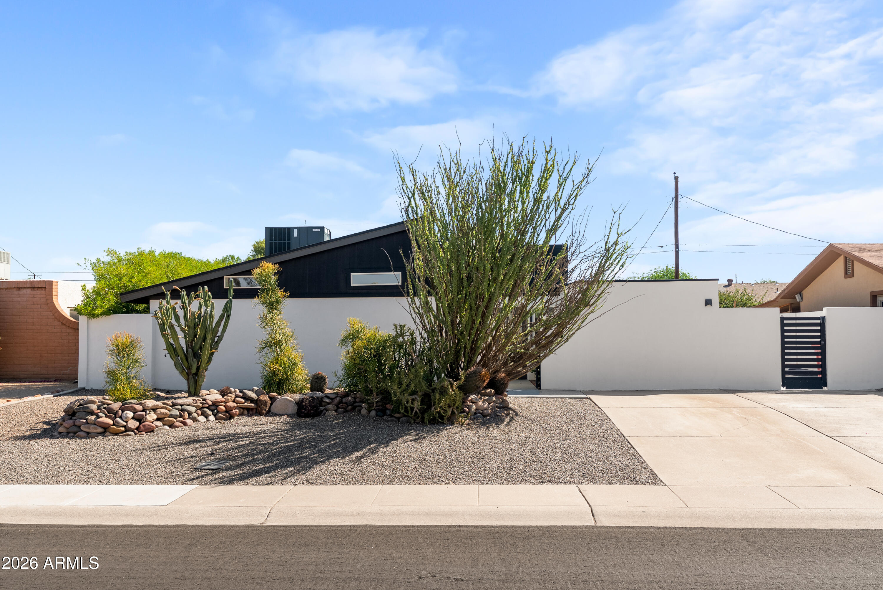 Modern home with a dark sloped roof, white walls, and desert landscaping behind a gated driveway.