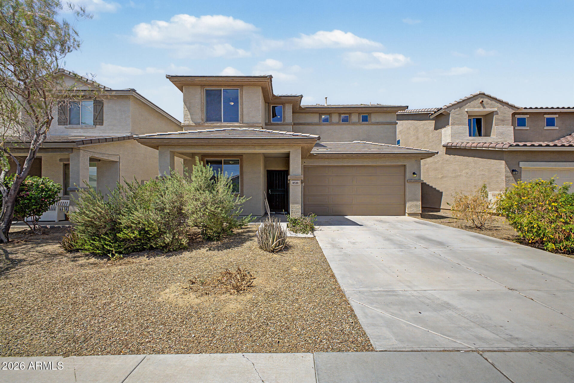 Two-story beige stucco home with a tiled roof, double garage, and desert landscaping.