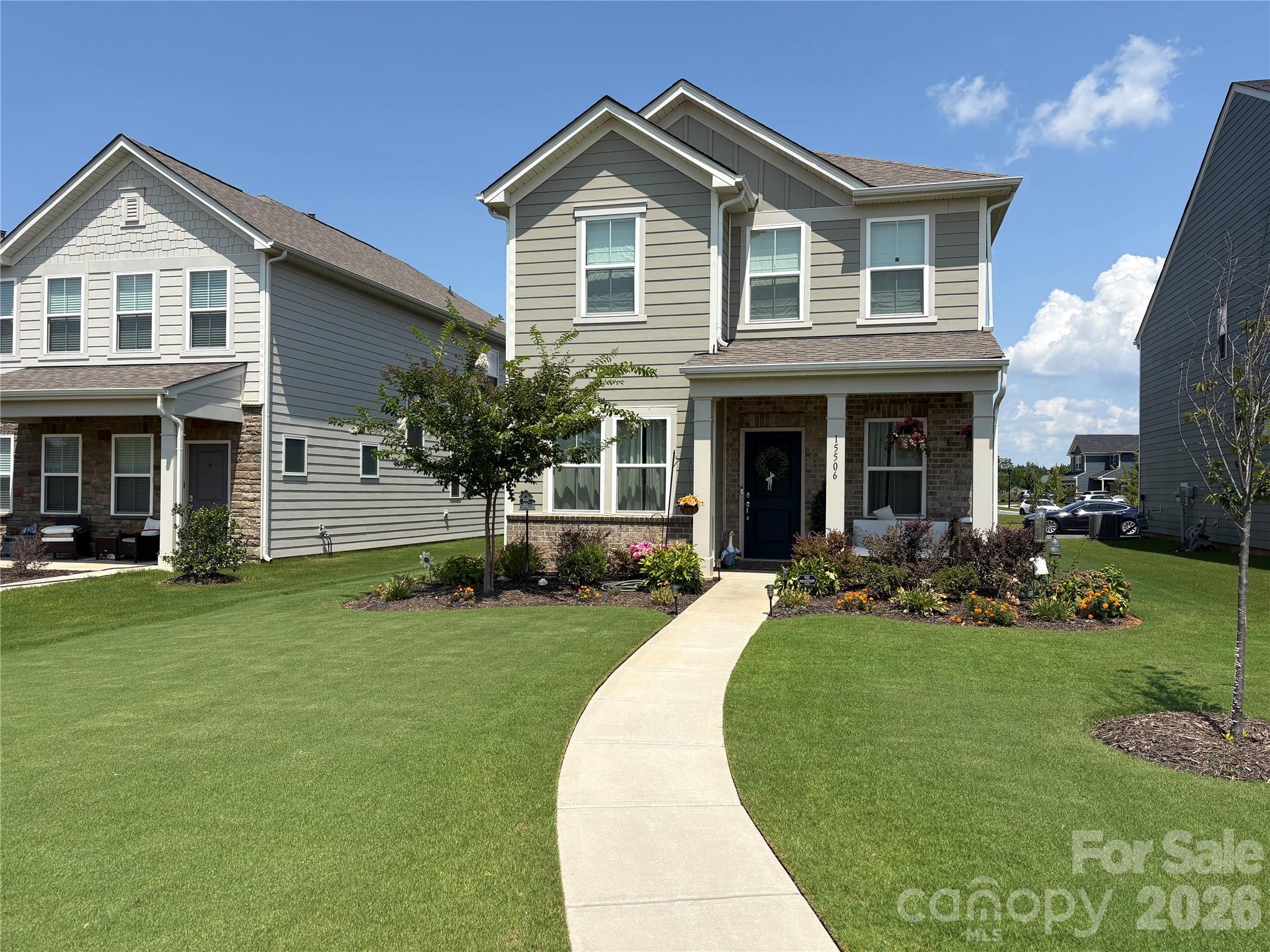 Two-story gray house with a brick porch, landscaped flower beds, and a curved walkway to the entrance.