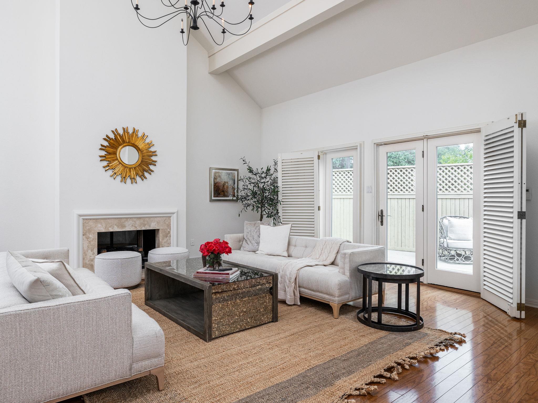 Living room with a fireplace, vaulted ceiling, pale sofas, and open shutters beside French doors.