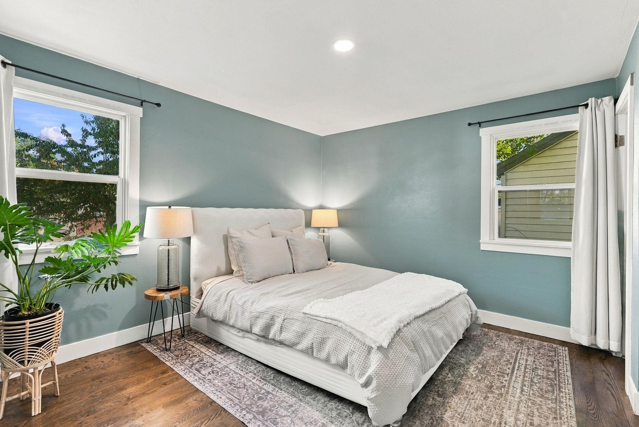 Staged bedroom with a white bed, blue-green walls, two windows, and a potted plant.