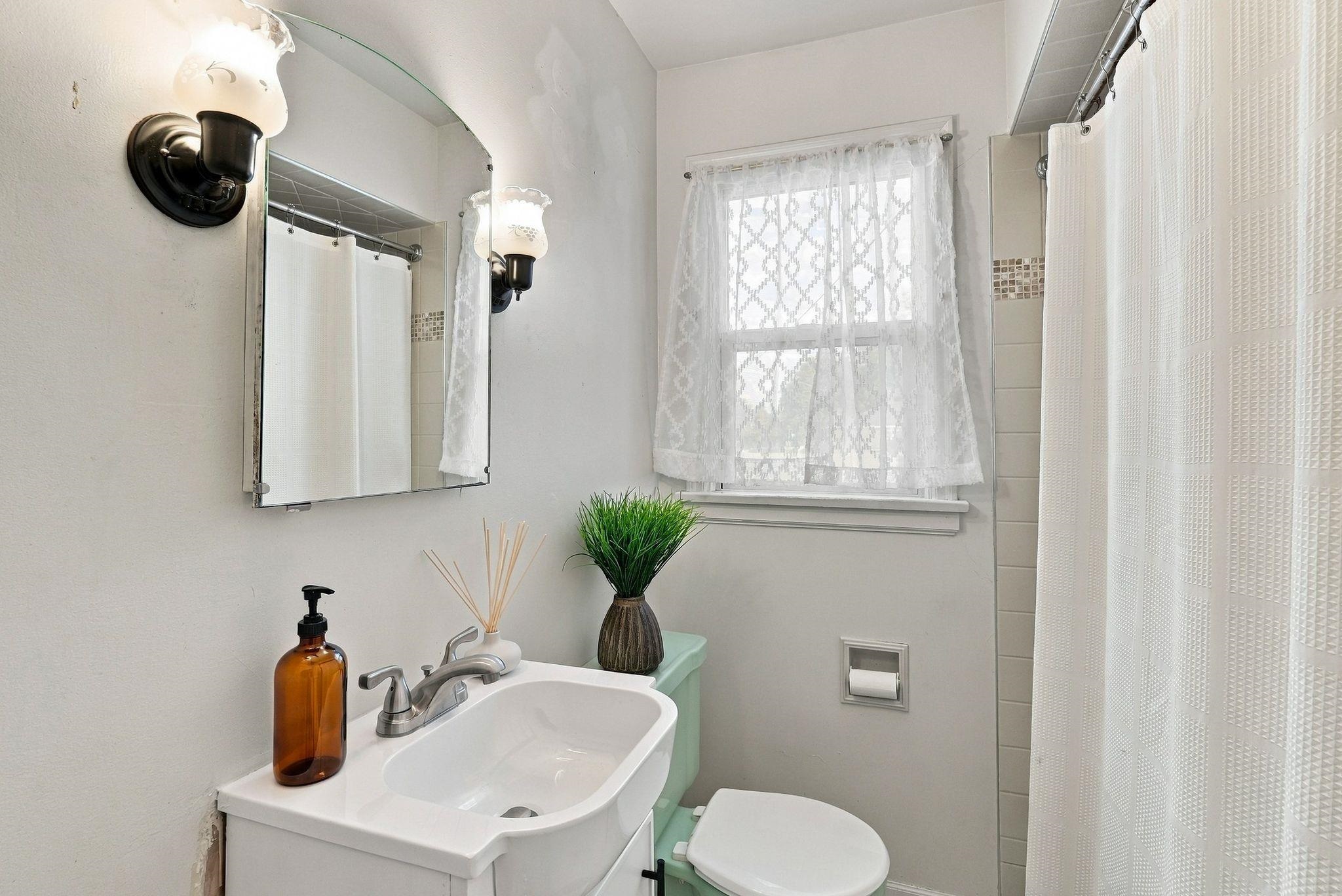 Bathroom with a white vanity, toilet, and curtained shower beside a lace-covered window.