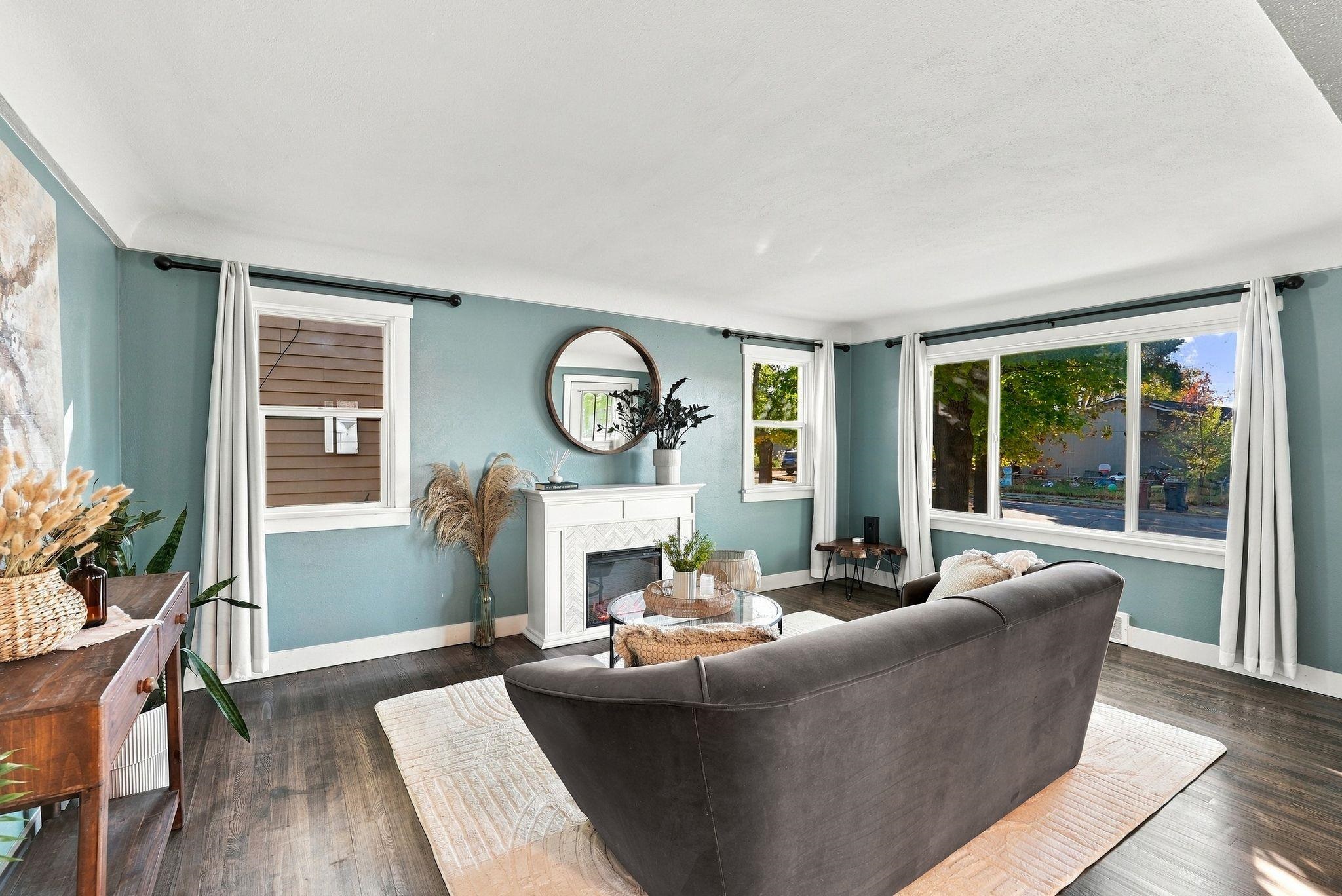 Staged living room with a white fireplace, gray sofa, and large windows.