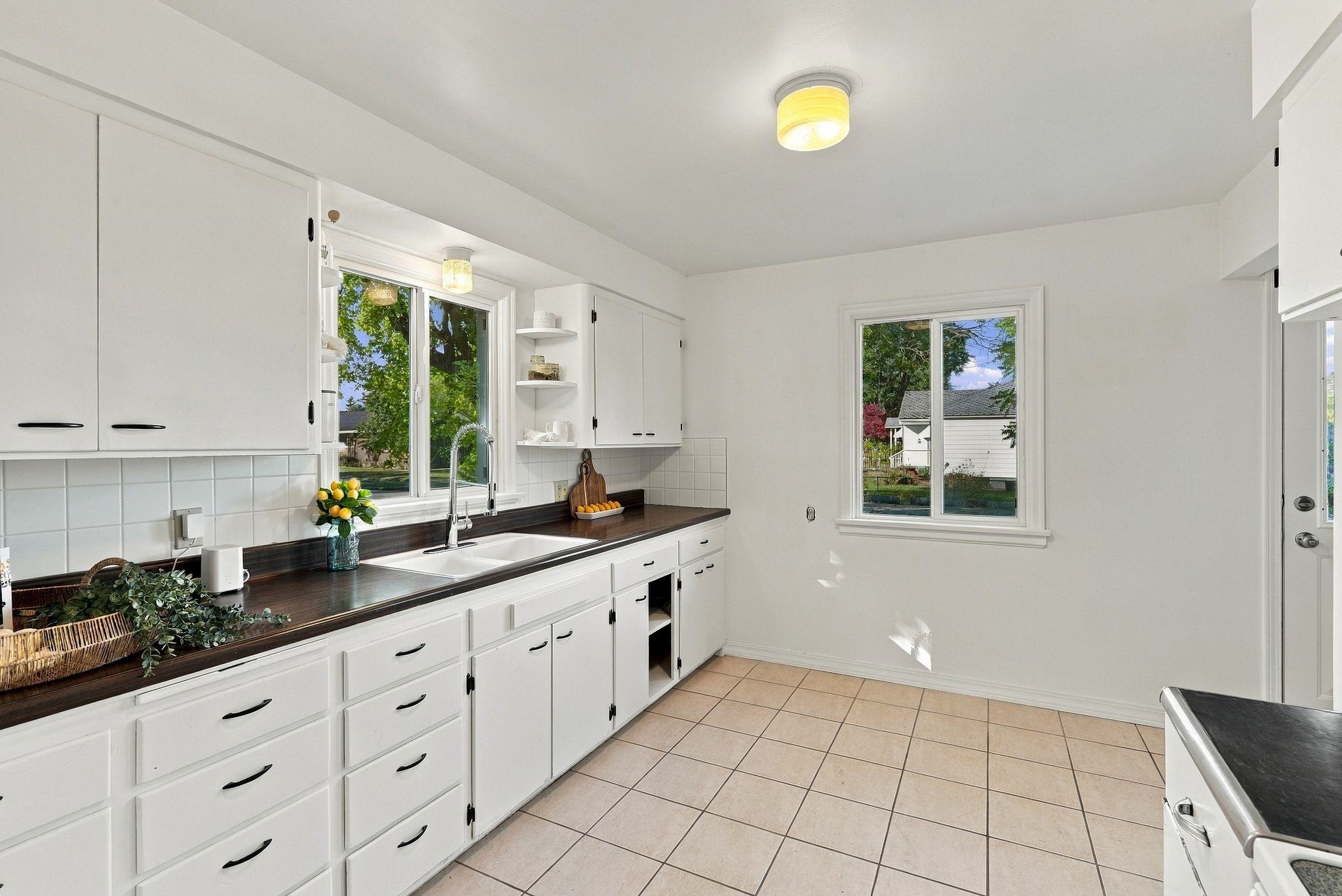 Kitchen with white cabinets, dark countertops, and two windows overlooking the yard.