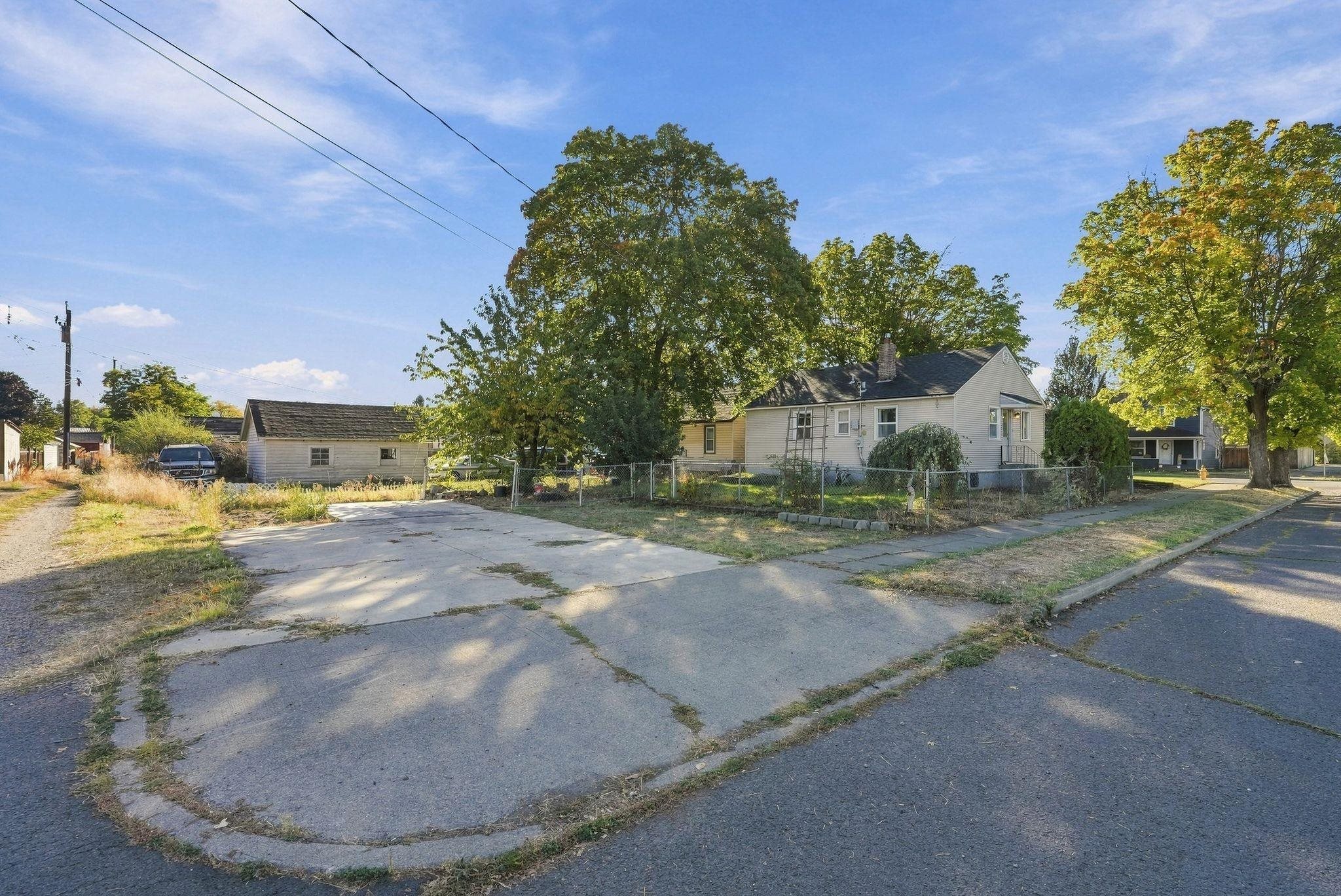 Single-story house behind a chain-link fence, with a broad concrete driveway and mature trees.