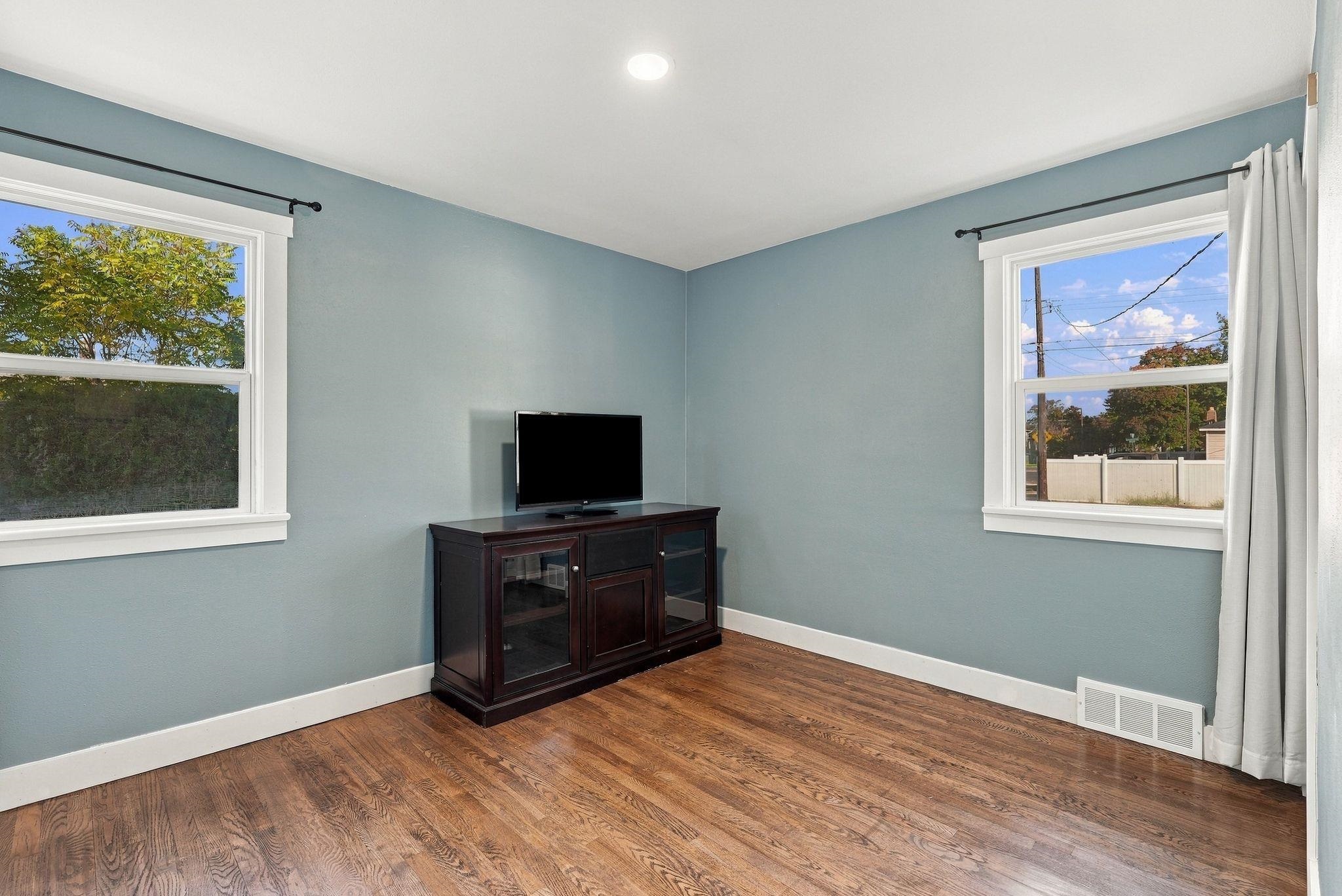 Blue-walled room with hardwood floors, two windows, and a television on a dark wood cabinet.