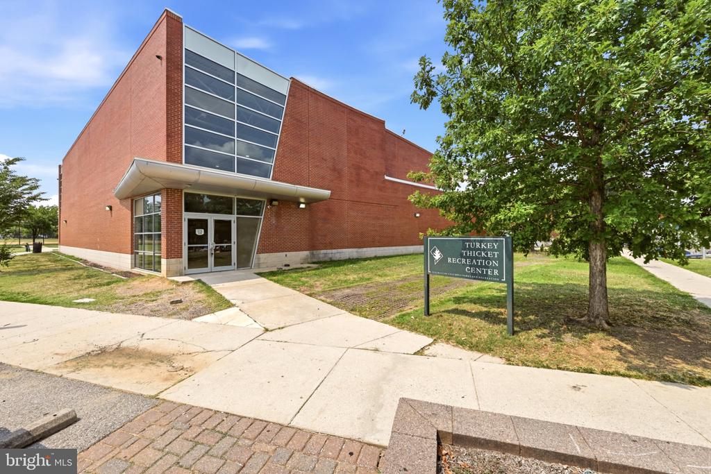 Turkey Thicket Recreation Center exterior with red-brick walls, a glass entrance, and a sign beneath a leafy tree.