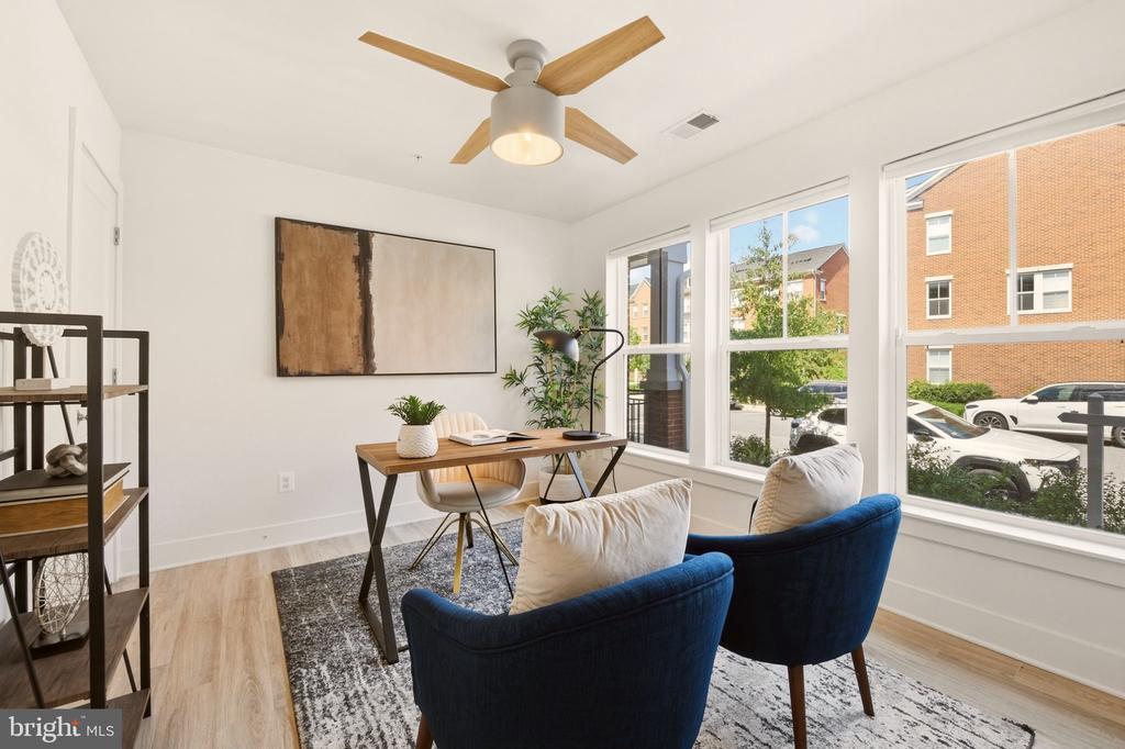 Staged home office with a wood desk, blue chairs, and large street-facing windows.