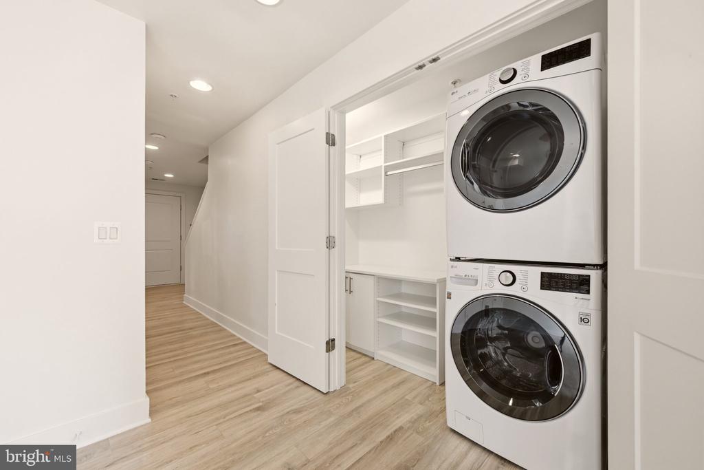 Laundry room with stacked washer and dryer, built-in shelving, and open doors to a wood-floored hallway.