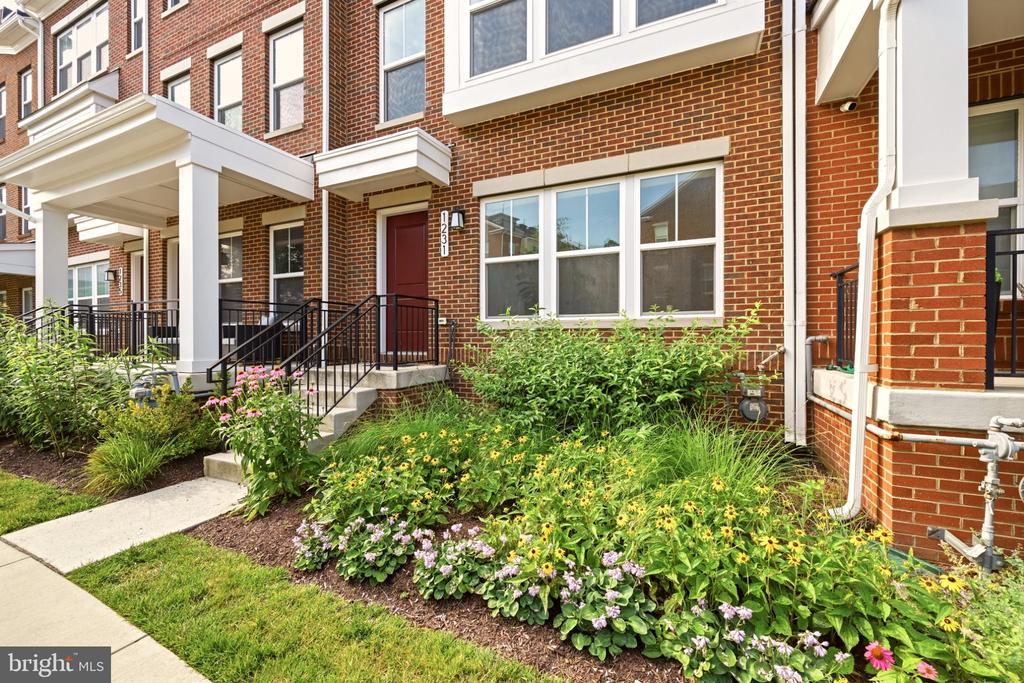 Red-brick townhouse entrance with white trim, black railings, and colorful flower beds.