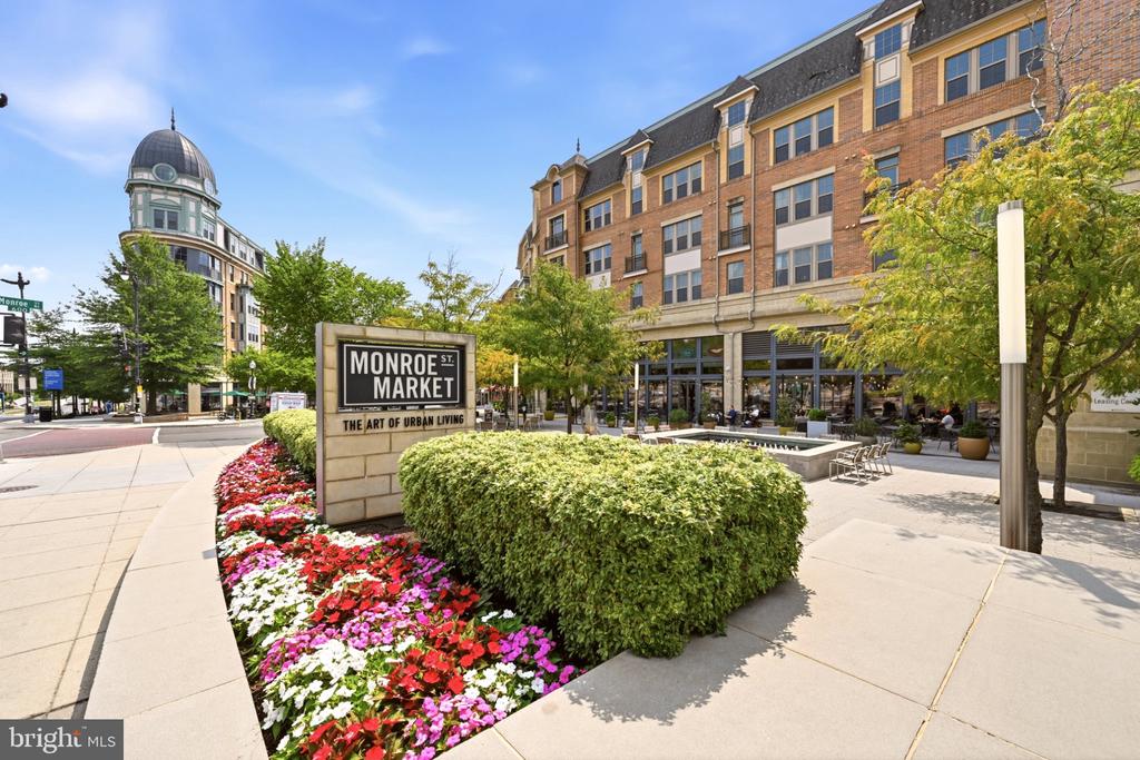 Monroe St. Market streetscape with brick buildings, outdoor seating, flower beds, and a landscaped plaza.