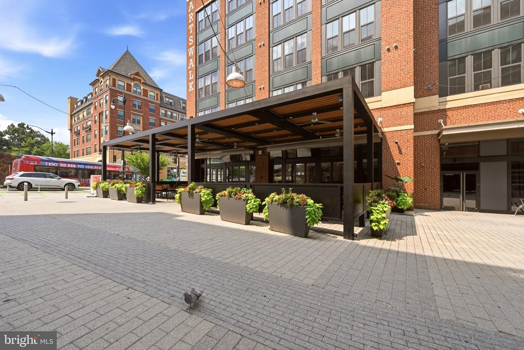 Brick and glass building with a covered patio and planters beside a paved plaza.