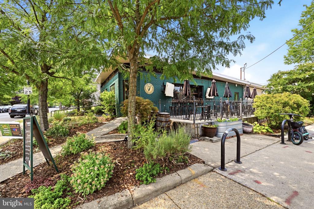 Teal commercial building with a fenced patio, outdoor tables, umbrellas, and leafy landscaping.