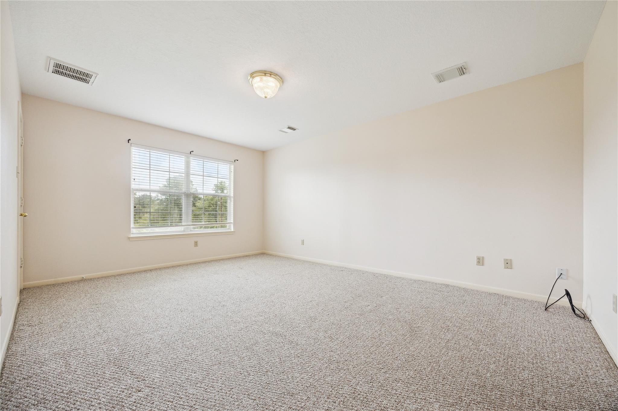 Empty carpeted room with beige walls, a window with blinds, and a ceiling light.