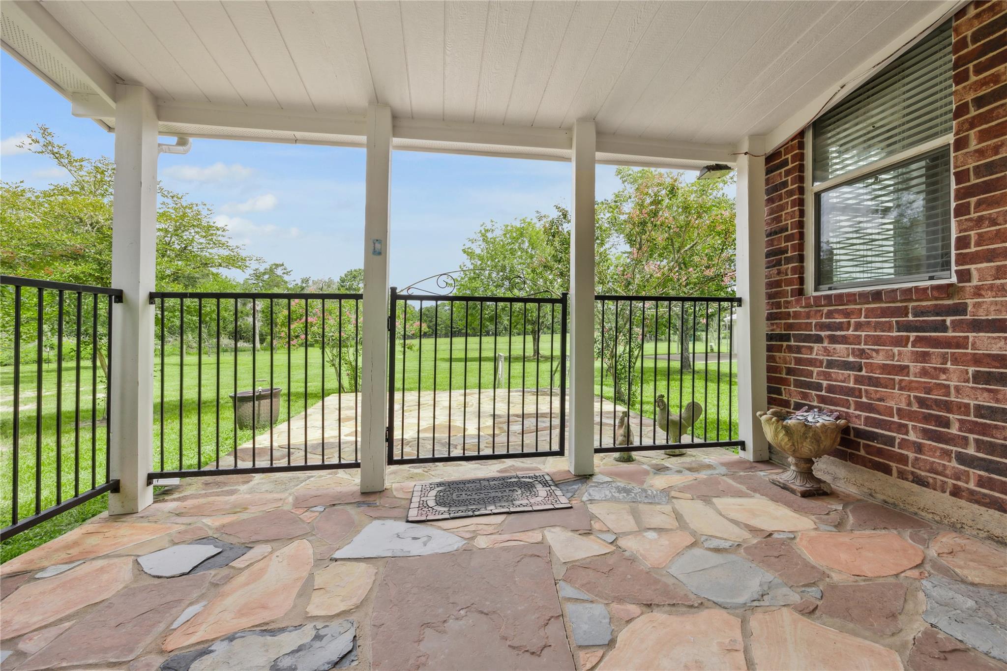 Covered flagstone porch with black railing overlooking a leafy lawn and trees.
