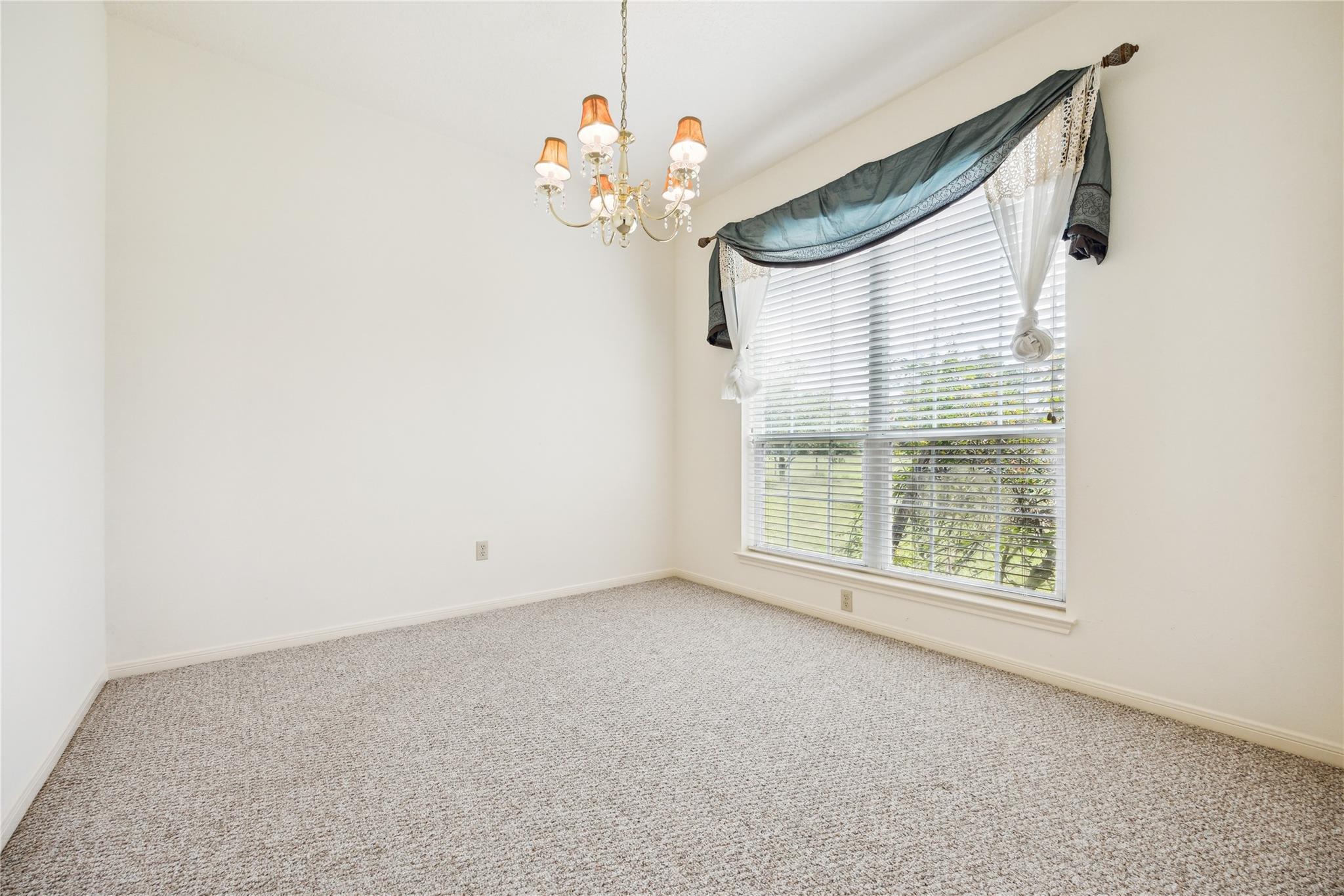 Empty carpeted room with a large window, decorative curtains, and a chandelier.