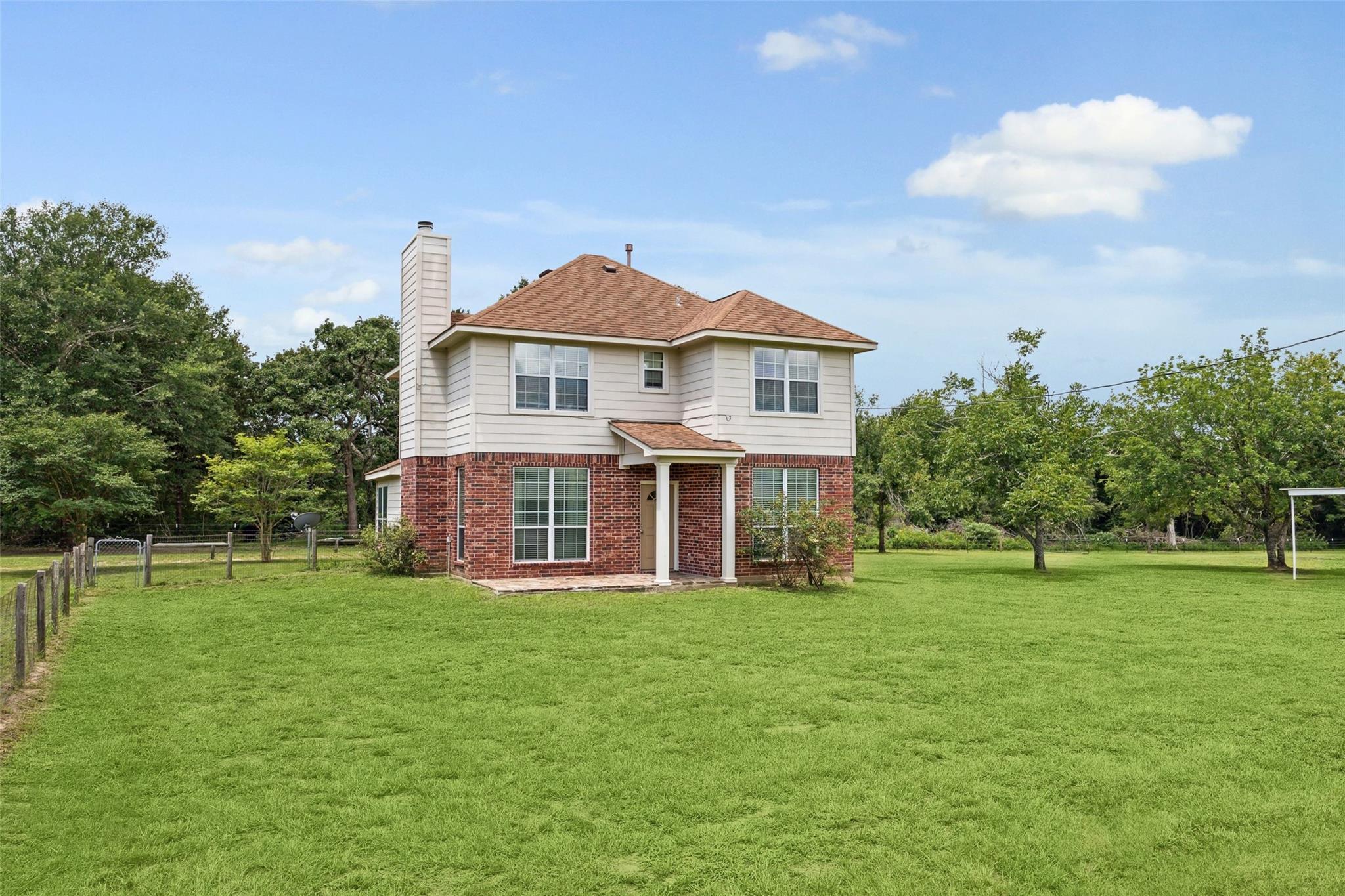 Two-story brick and siding house with a covered entry, broad lawn, mature trees, and a fenced yard.