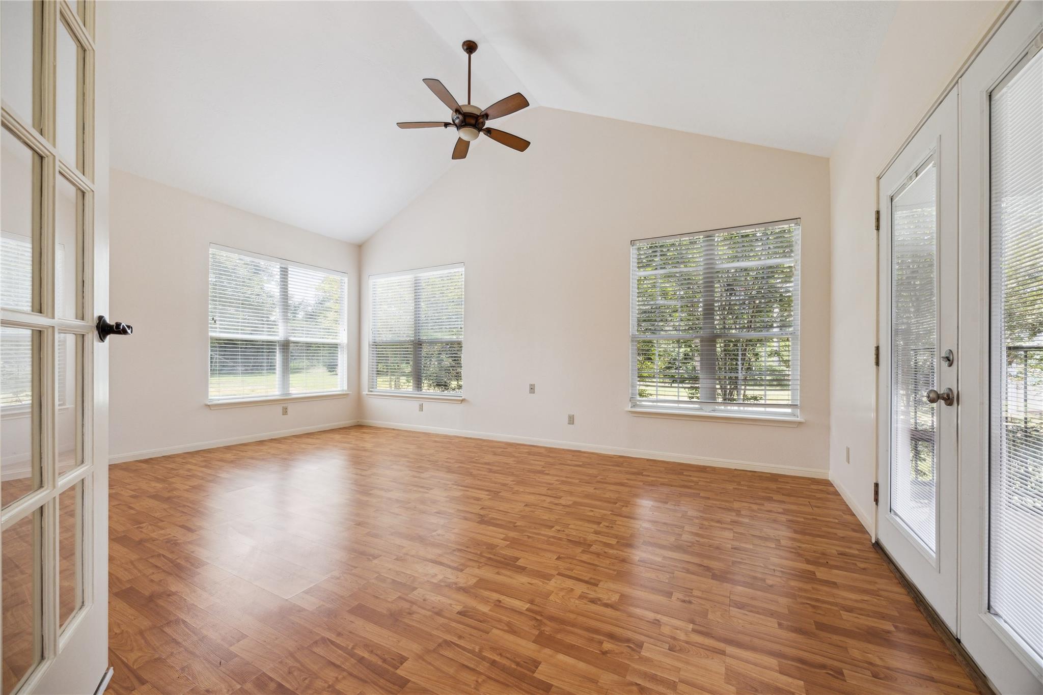 Empty room with wood flooring, a vaulted ceiling, ceiling fan, and large windows overlooking greenery.