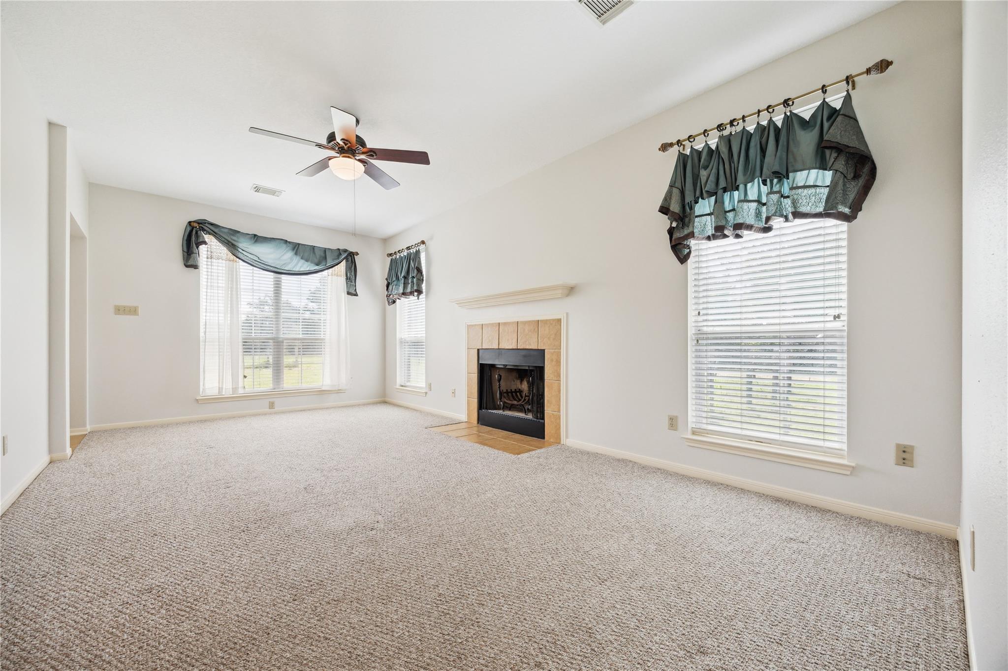 Empty living room with a tiled fireplace, ceiling fan, and windows with teal valances.