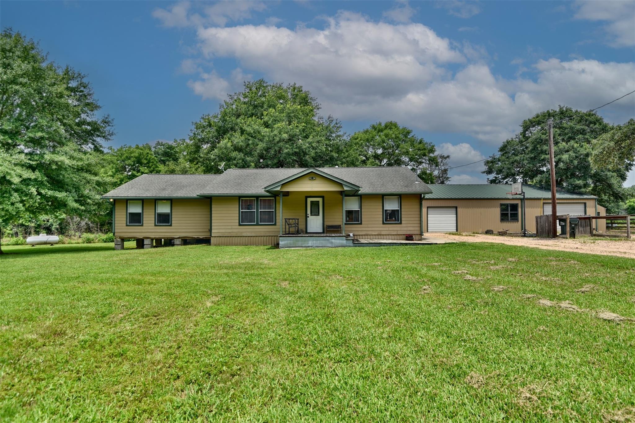 Single-story tan home with an attached metal-sided garage, broad green lawn, and mature trees.