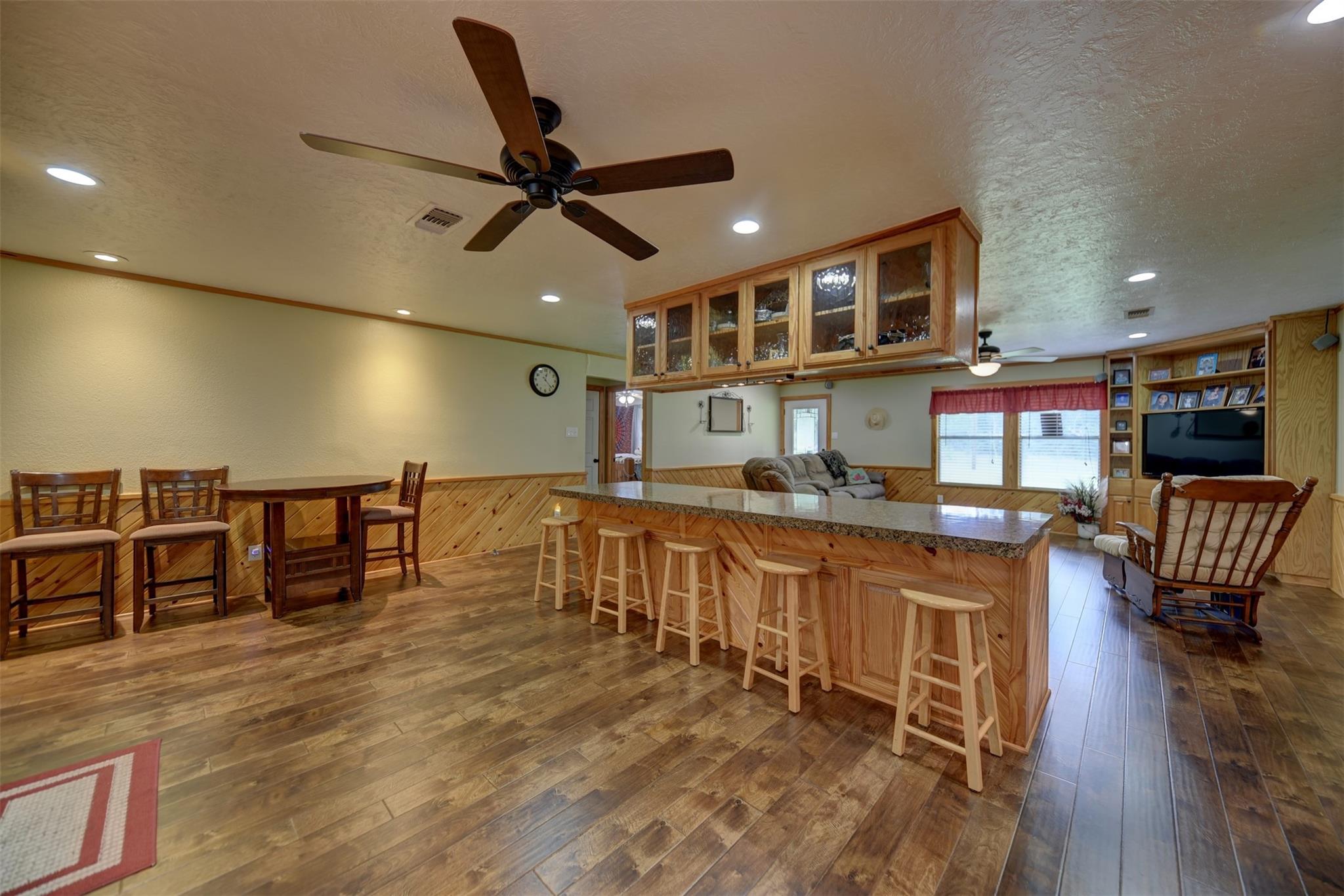 Open-plan kitchen and living area with a long granite island, wooden bar stools, and glass-front cabinets.