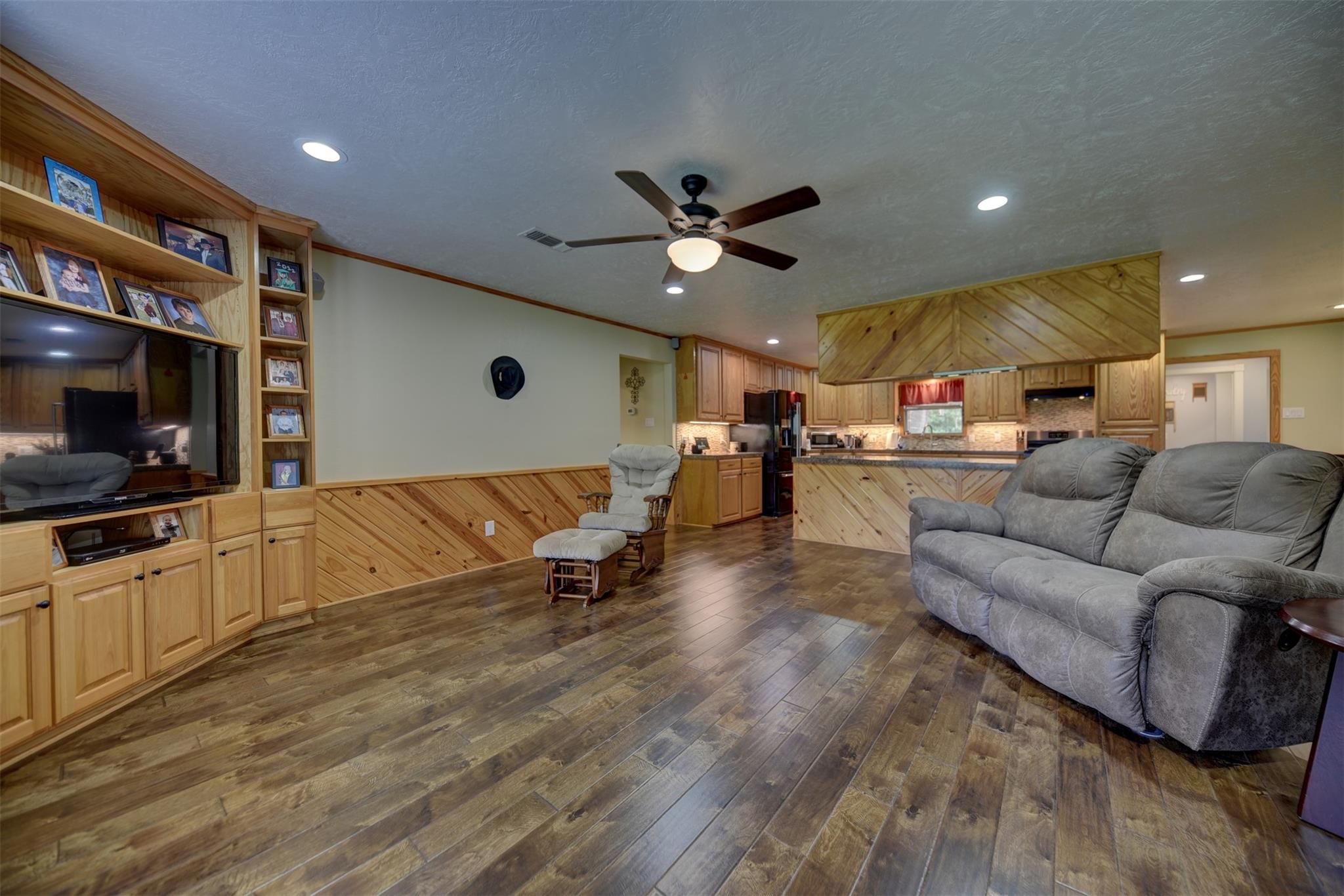 Open-plan living room and kitchen with wood paneling, built-in TV cabinetry, and gray reclining sofas.