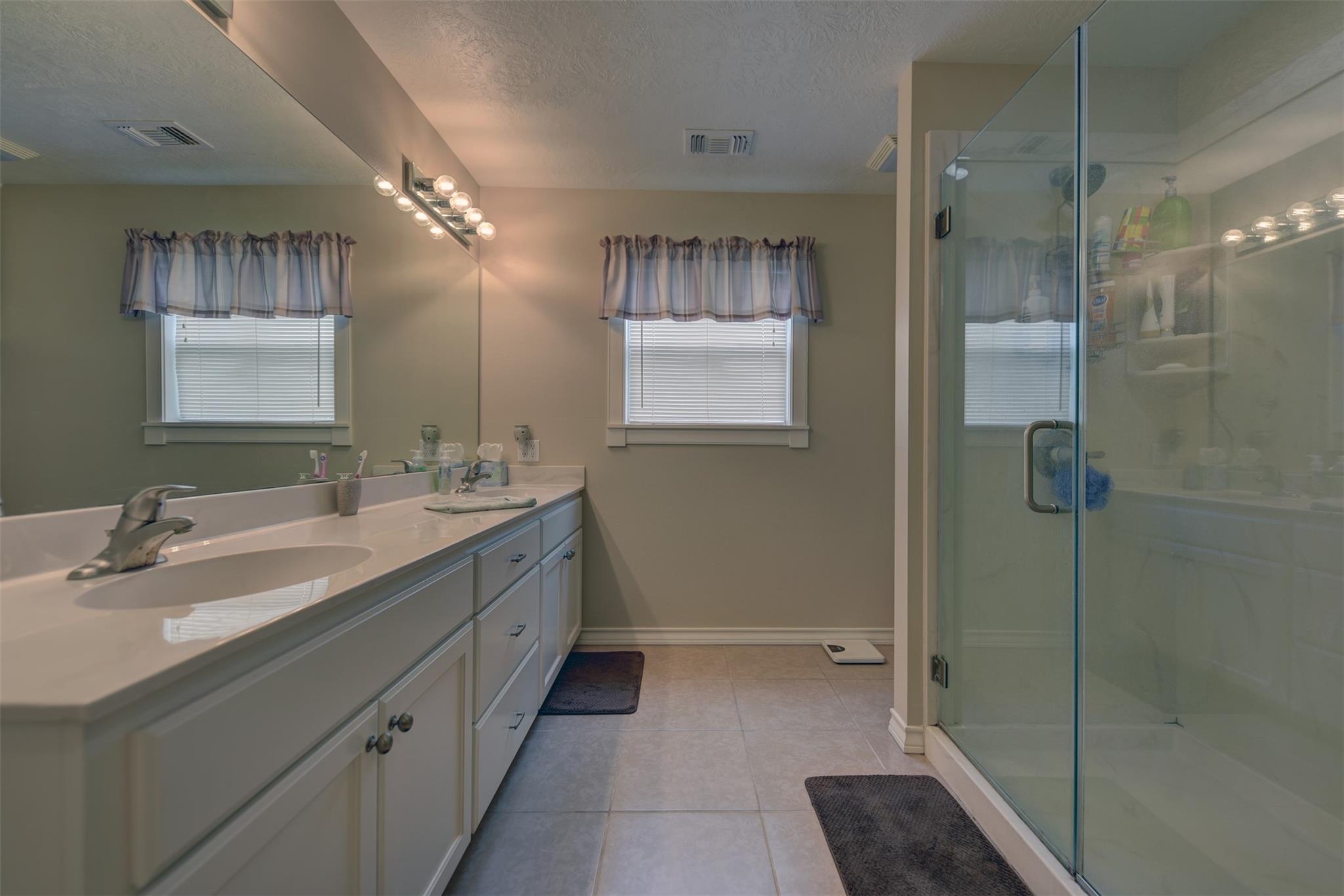 Bathroom with a long double-sink vanity, two windows, and a glass-enclosed shower.
