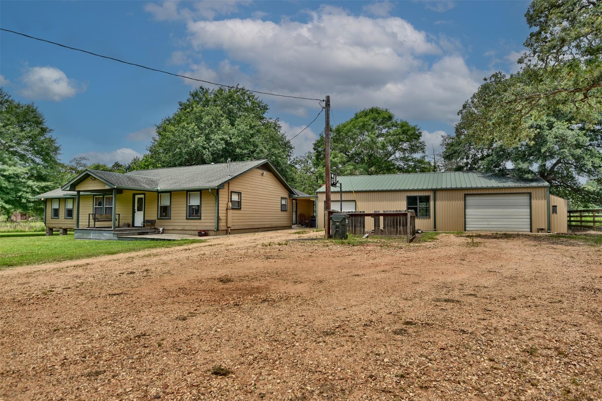 Tan single-story house with a covered porch beside a large metal outbuilding and gravel driveway.