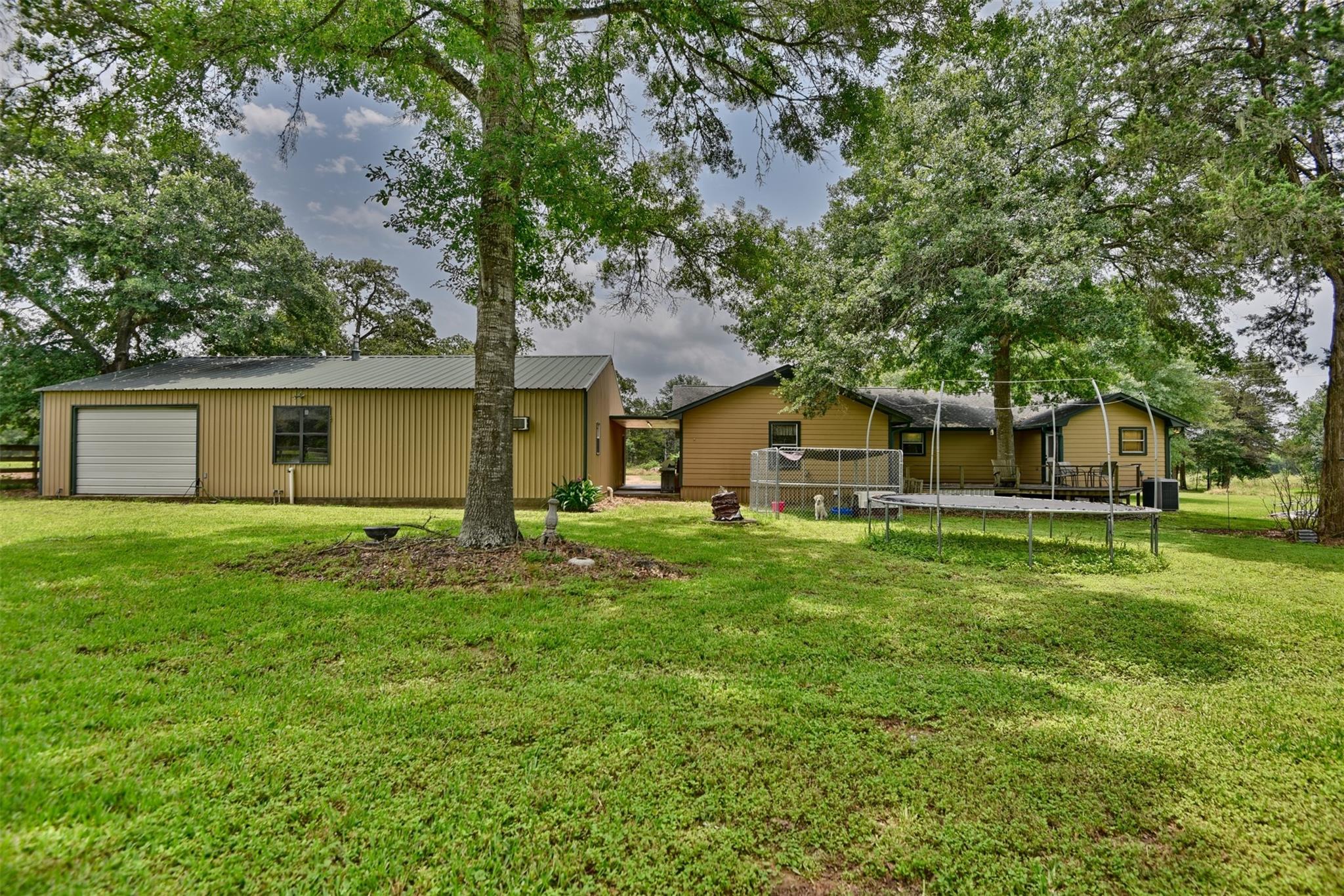 Single-story home and detached garage beneath mature trees, with a spacious lawn and raised backyard deck.
