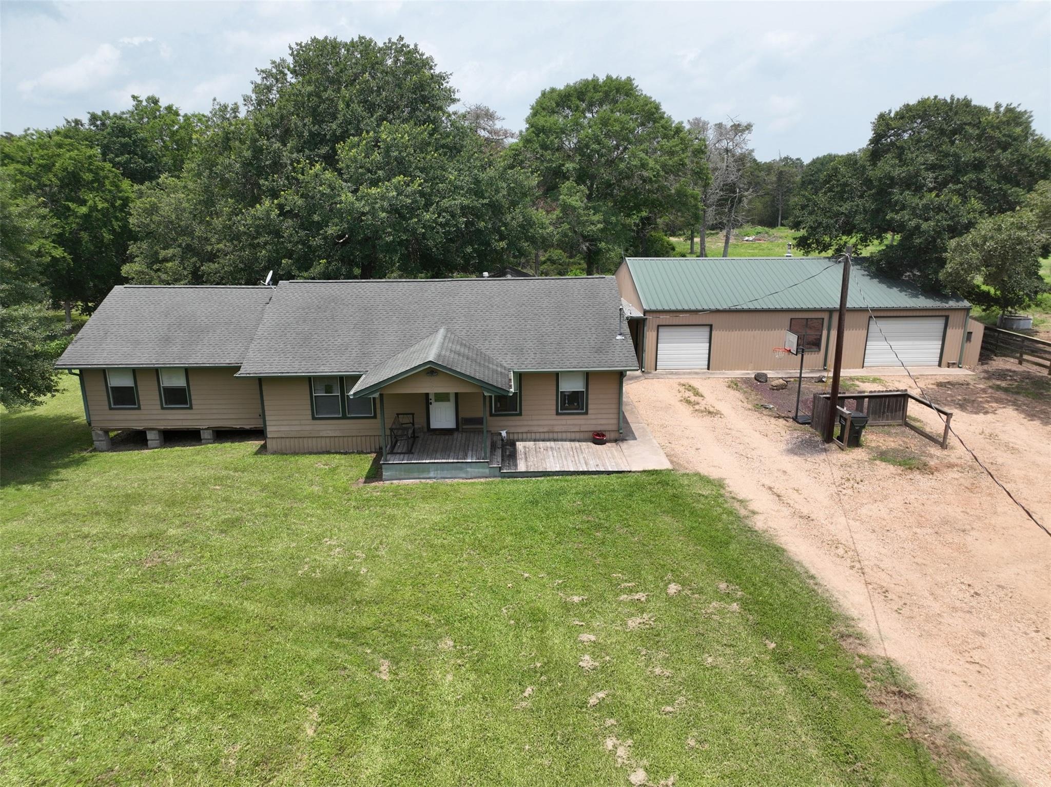 Aerial view of a tan house and detached metal outbuilding with a green roof, surrounded by lawn and trees.