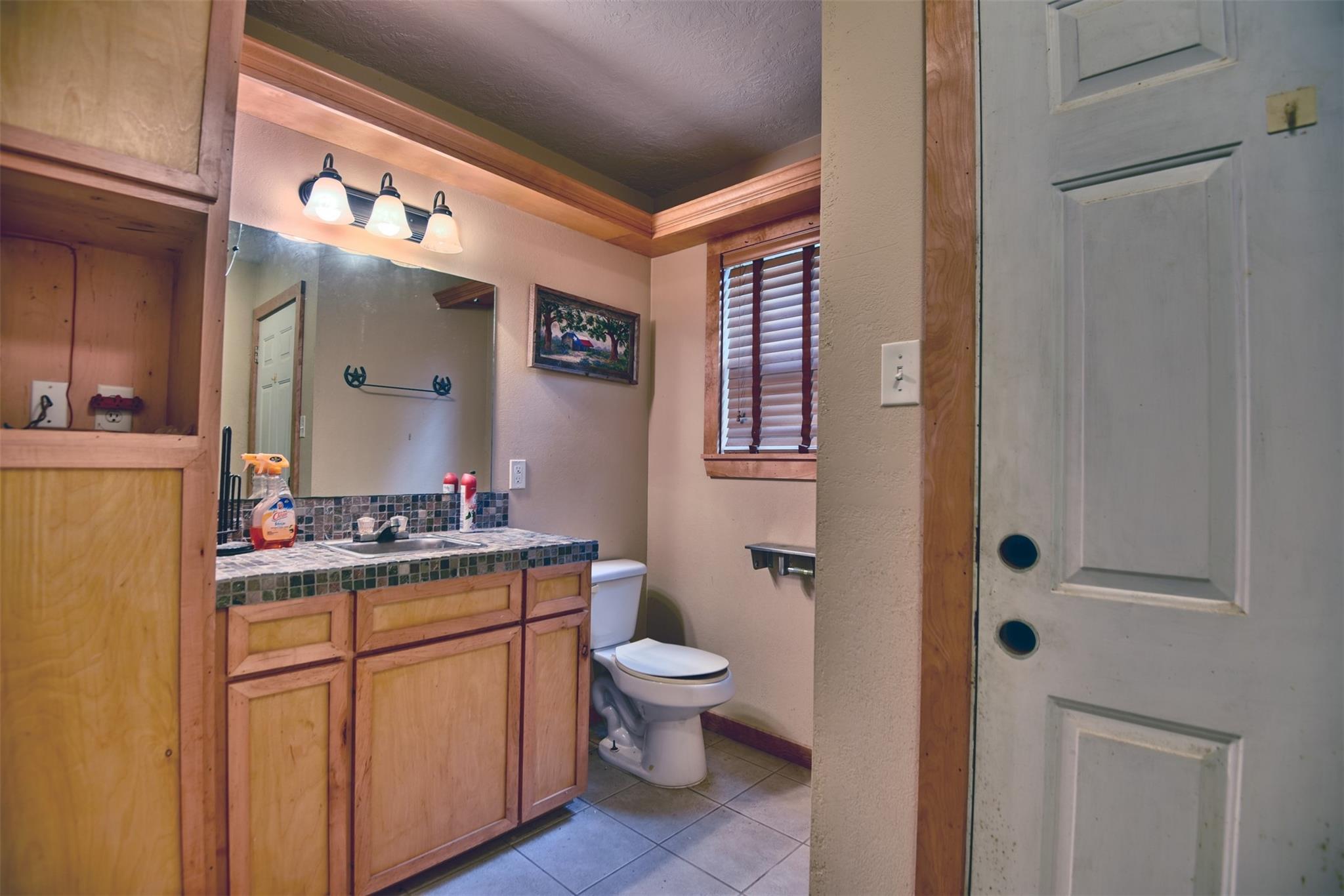Bathroom with a wood vanity, tiled counter, large mirror, and toilet beside a window with blinds.