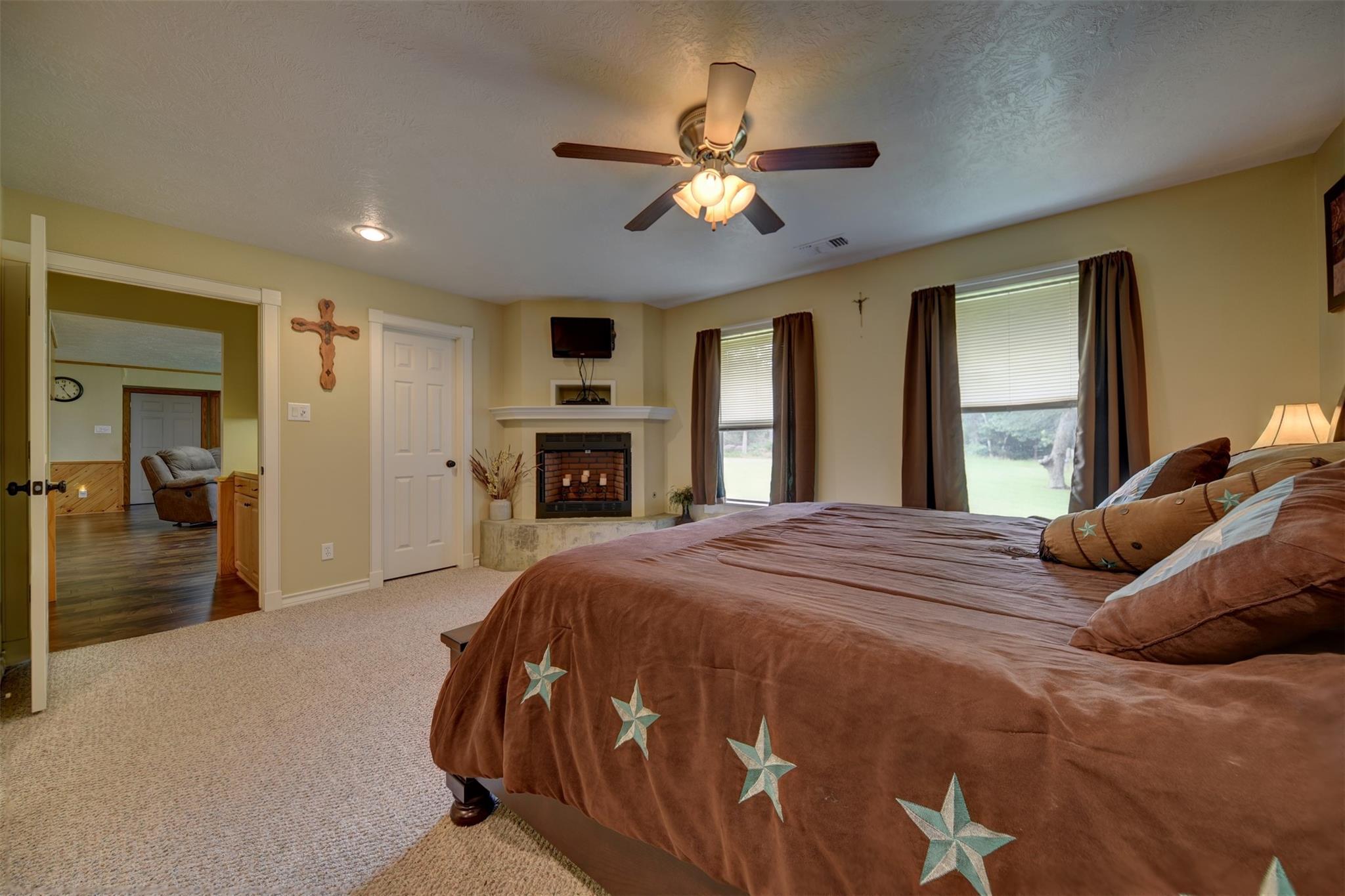 Furnished bedroom with a star-patterned bedspread, fireplace, ceiling fan, and windows overlooking a lawn.