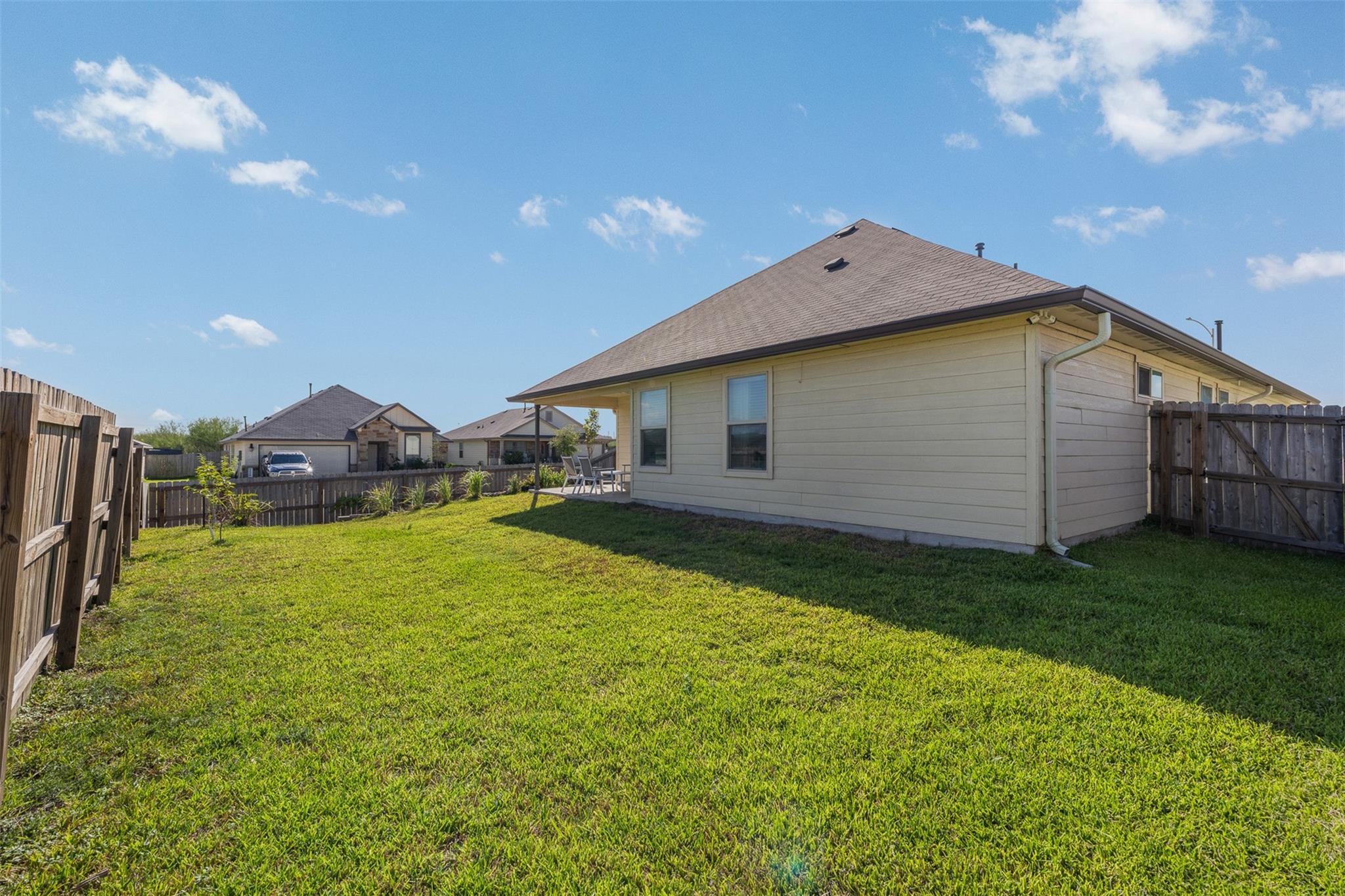 Home’s fenced backyard with a broad lawn, light siding, and a covered patio.