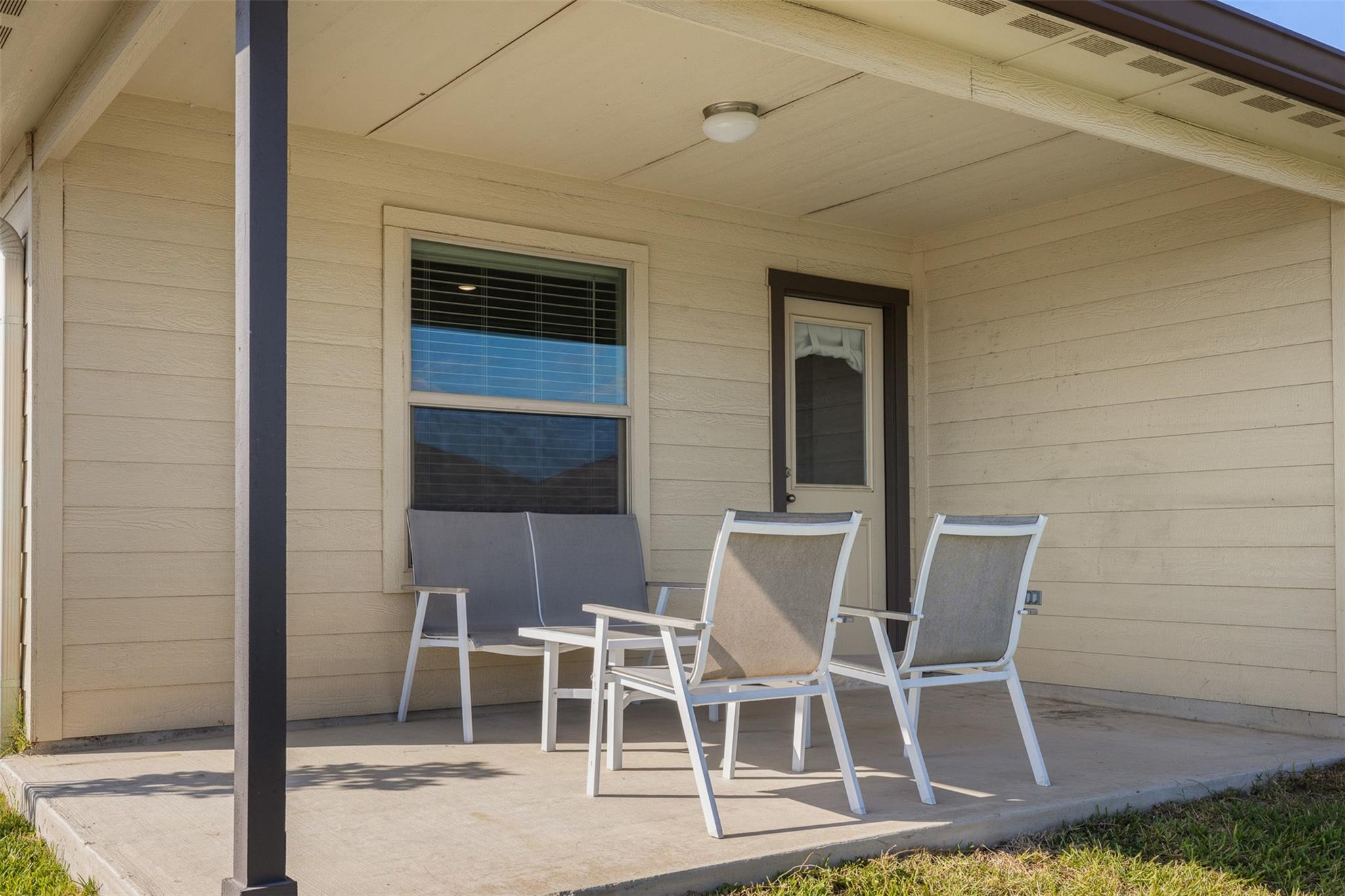 Covered porch with beige siding, a window, glass-panel door, bench, and two chairs.