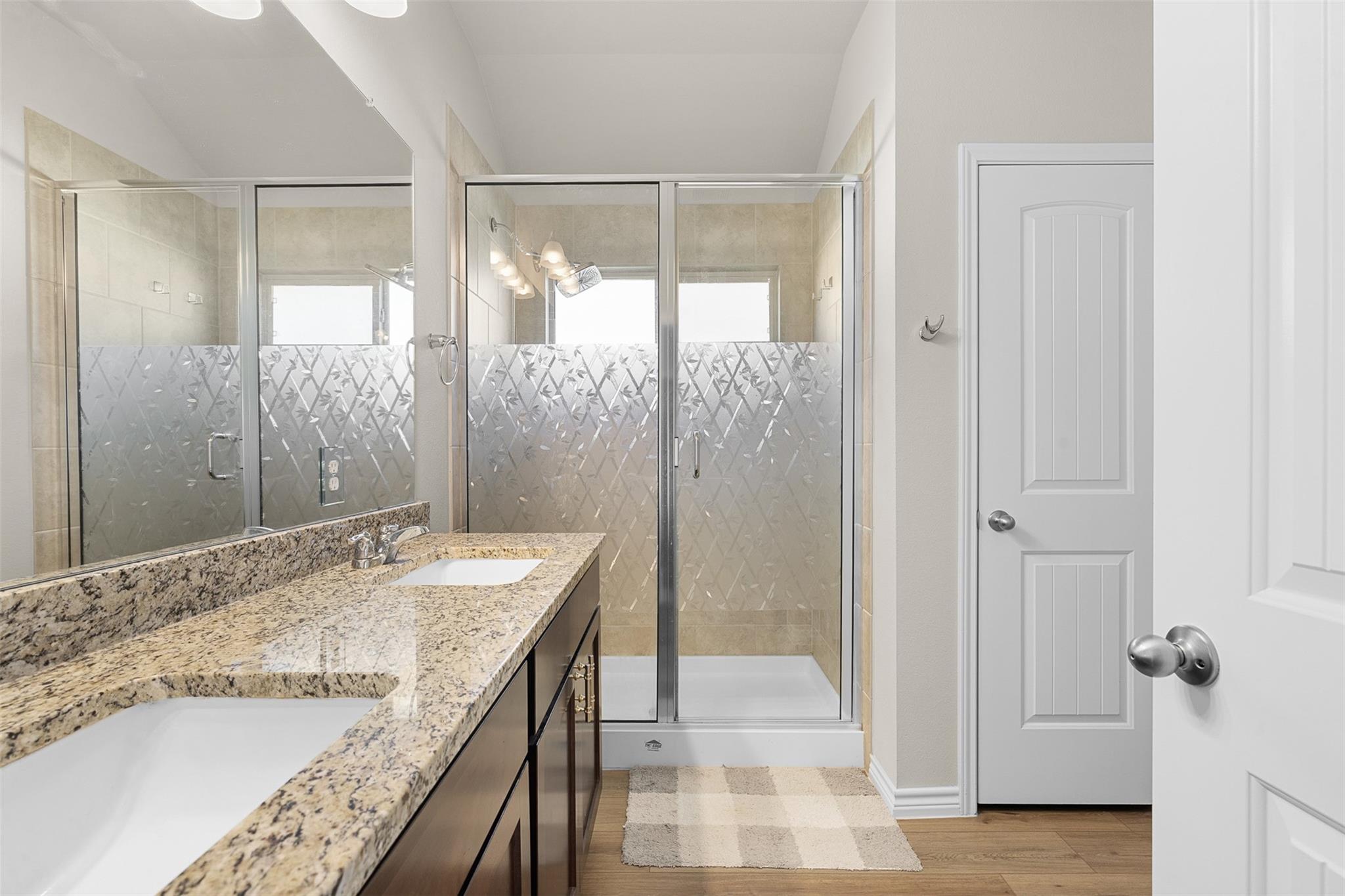 Bathroom with granite double vanity and glass shower featuring patterned privacy panels.