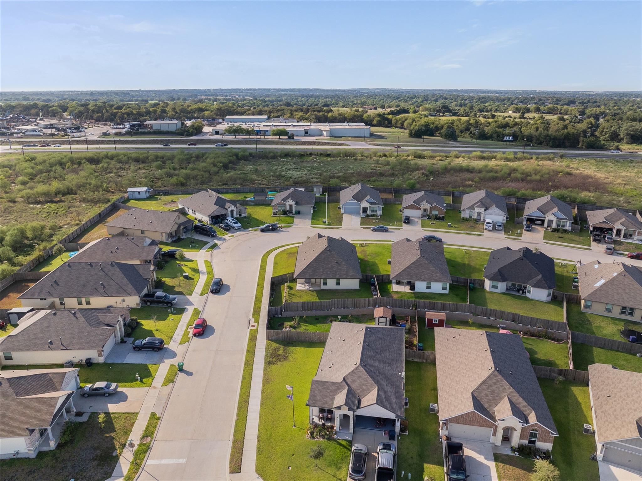 Aerial view of homes along a curving neighborhood street, with open land and a road beyond.