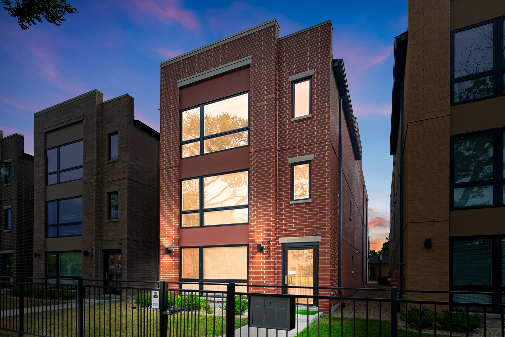 Red-brick townhouse with large windows and a fenced front yard at dusk.