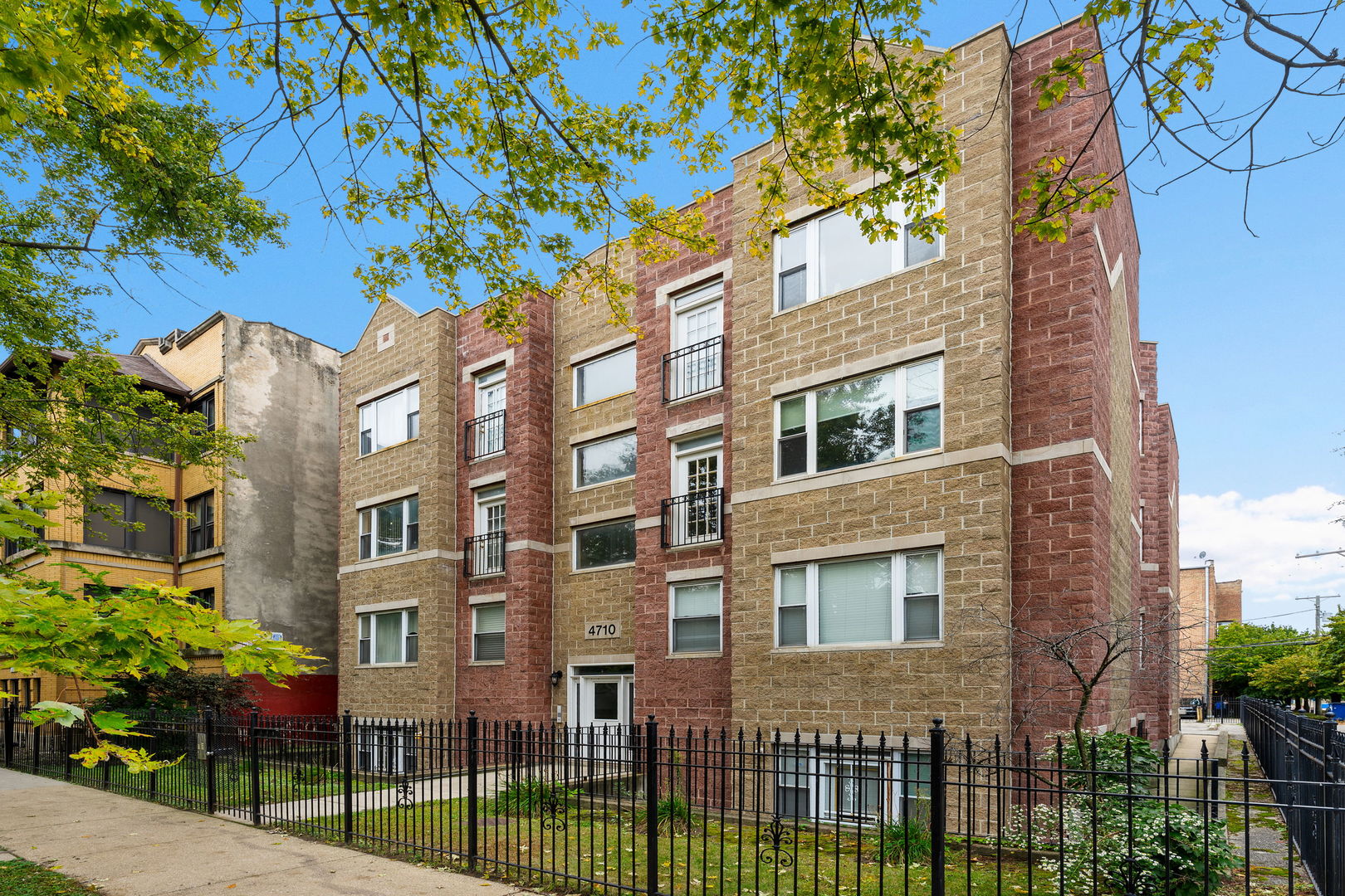 Multi-story tan and red brick apartment building behind a black metal fence and leafy trees.