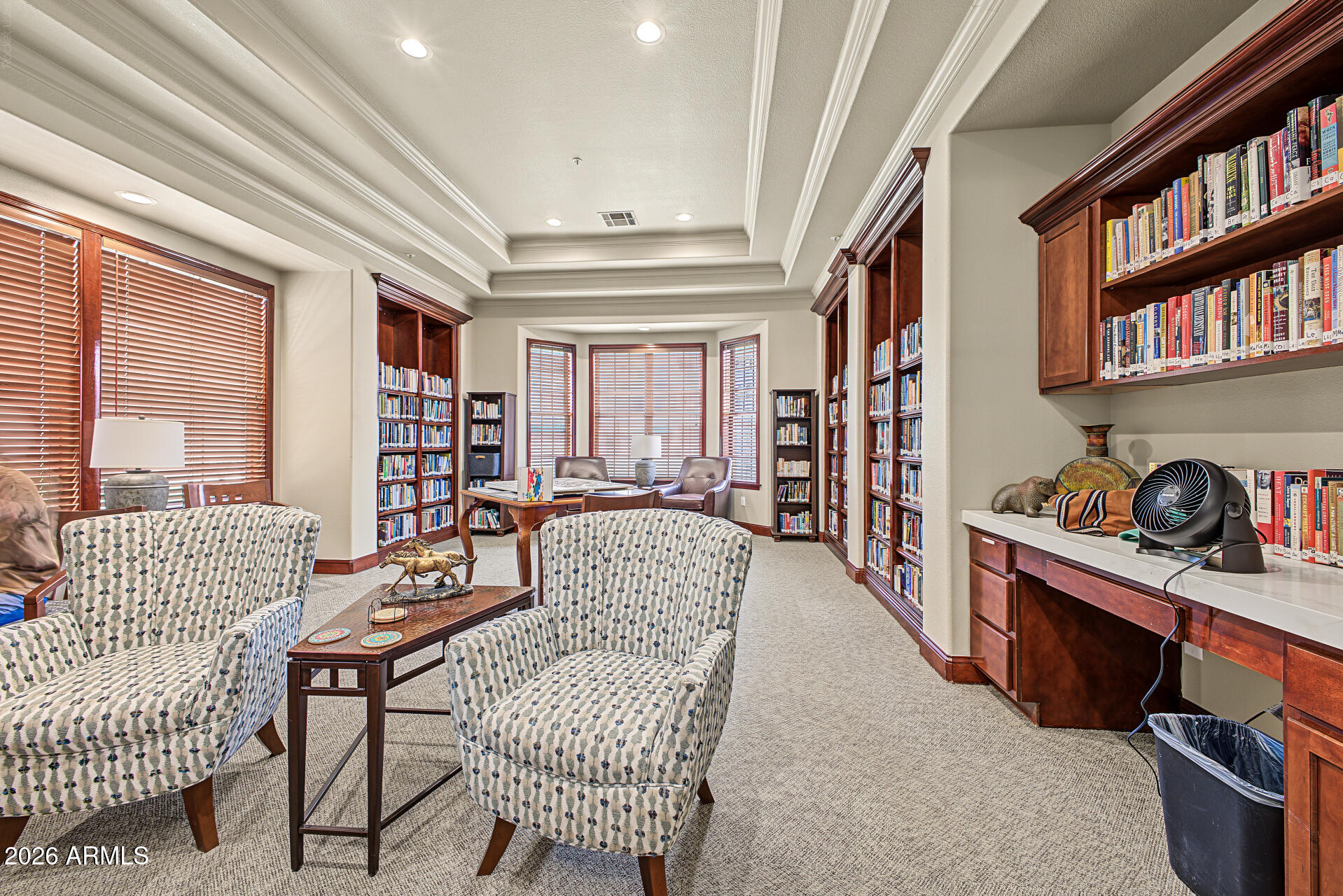 Furnished library with built-in bookcases, patterned armchairs, and large windows with wooden blinds.