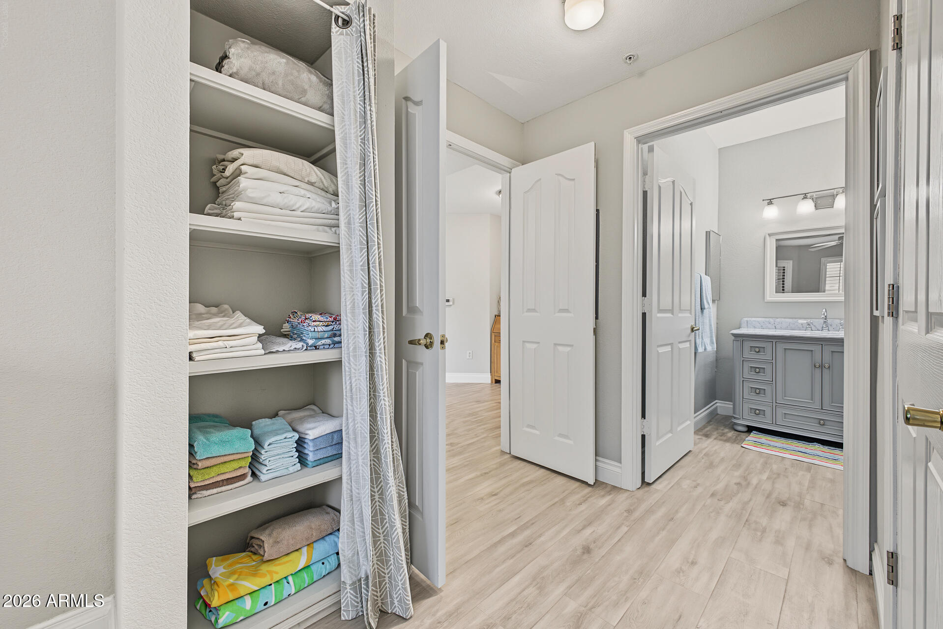 Linen shelves beside a bathroom with a gray vanity and light wood-look flooring.