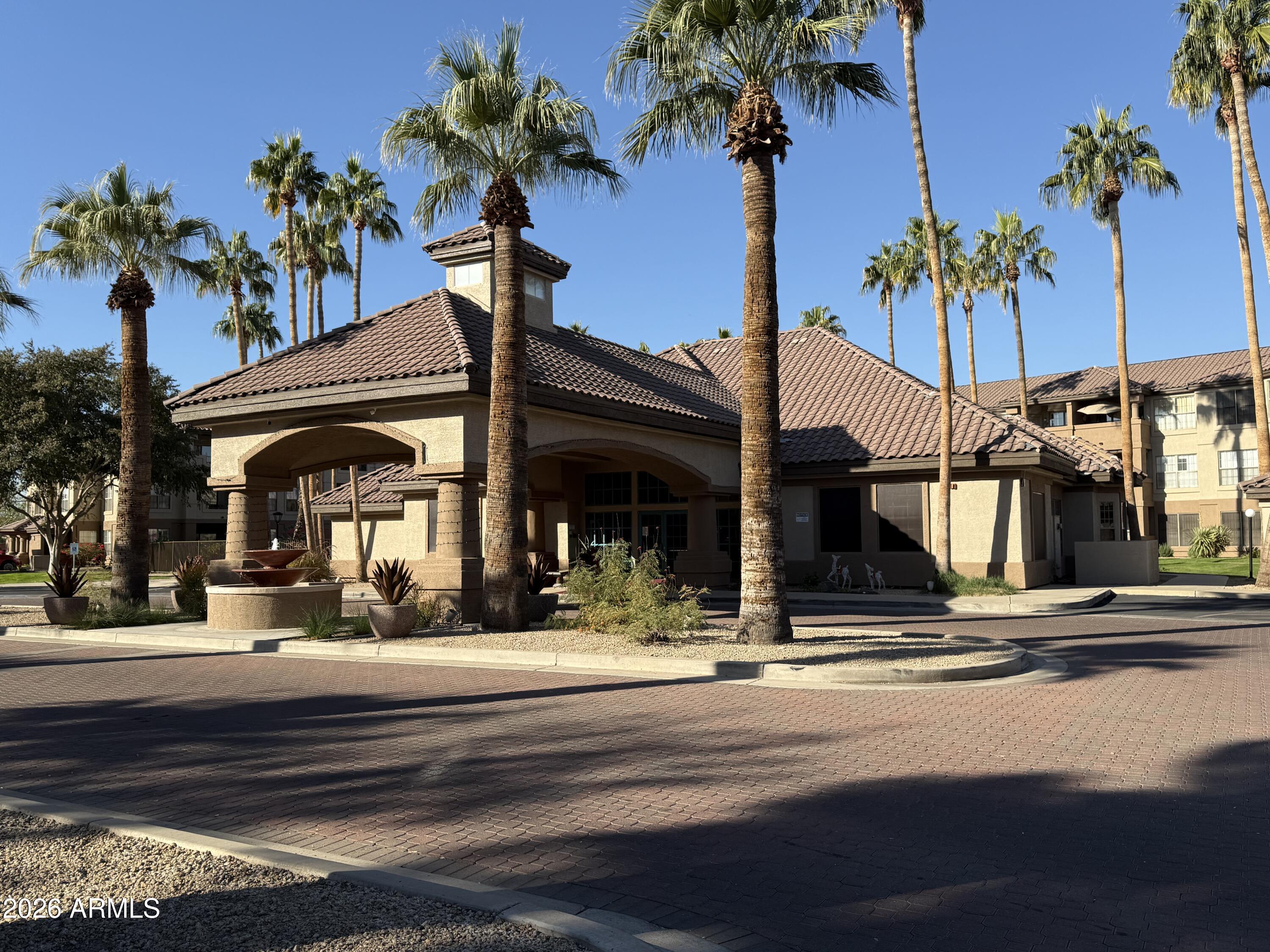 Residential complex entrance with a tiled roof, palm trees, and a courtyard fountain.